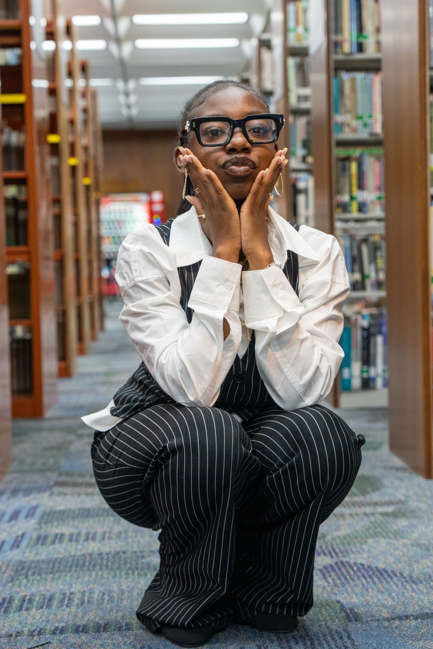 Young Black woman in glasses and pinstripe overalls crouching in library aisle, resting face in hands.
