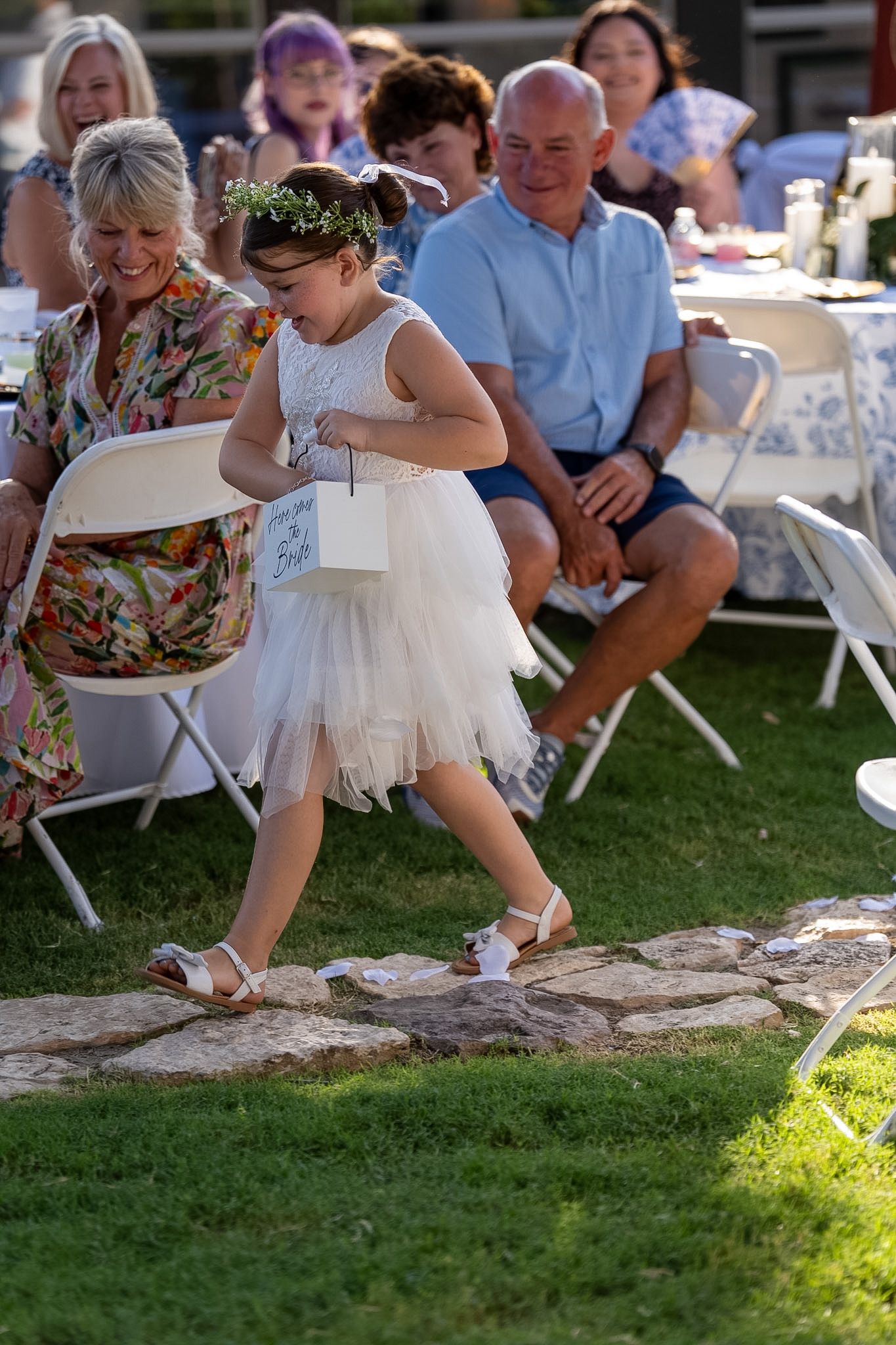 Young flower girl in white tutu dress walks stone path at outdoor wedding ceremony while guests watch.