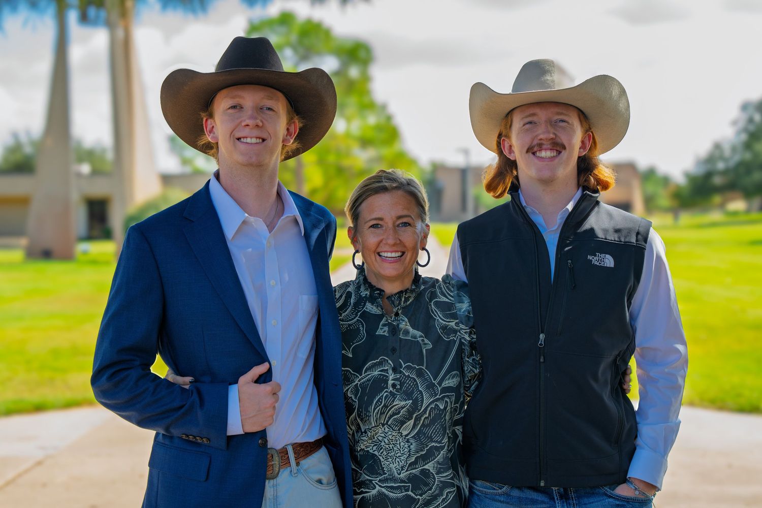 Three smiling people in cowboy hats pose outdoors on a sunny day, two men flanking a woman on a green campus.