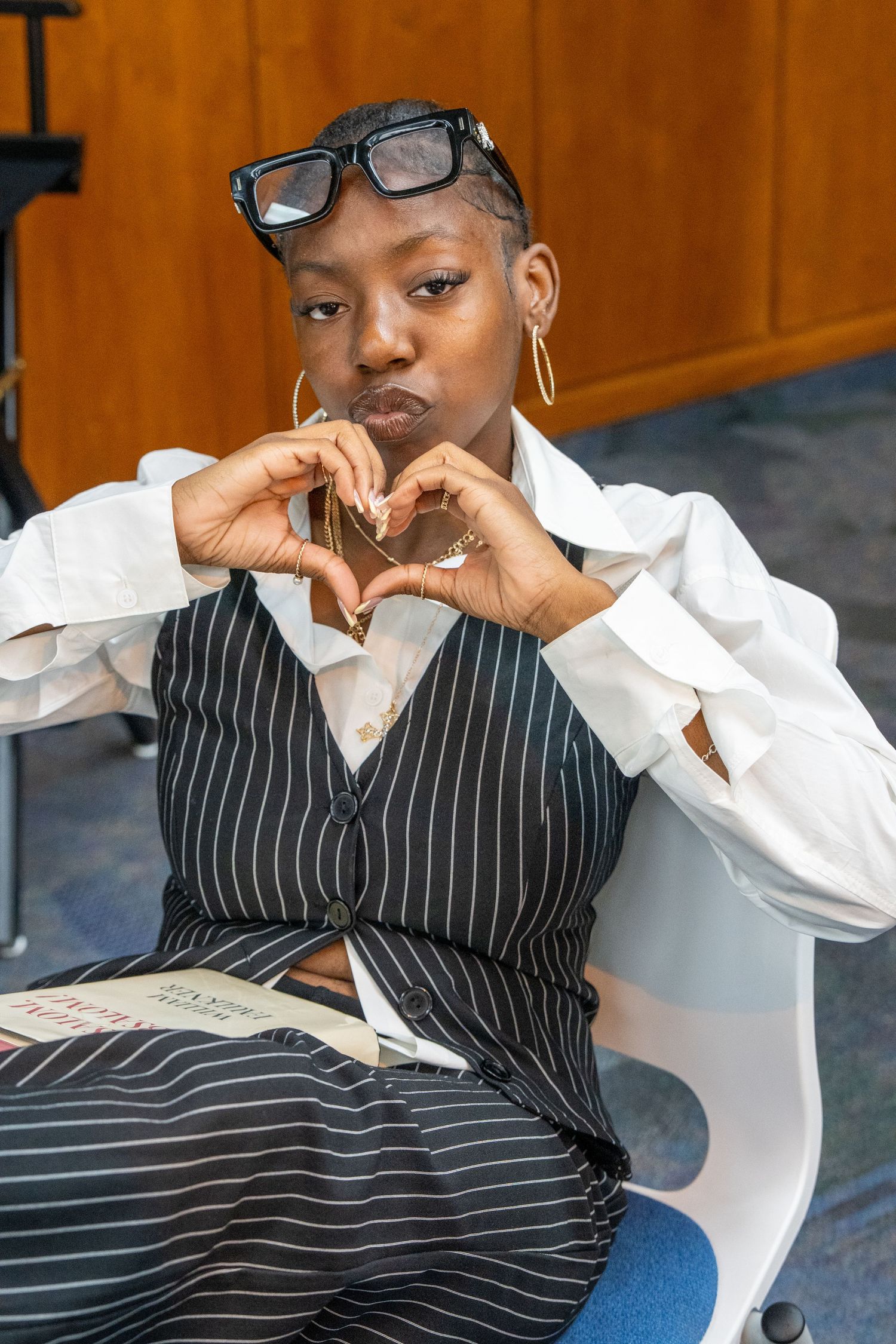 Young woman in pinstripe vest makes heart shape with hands while seated in white chair.
