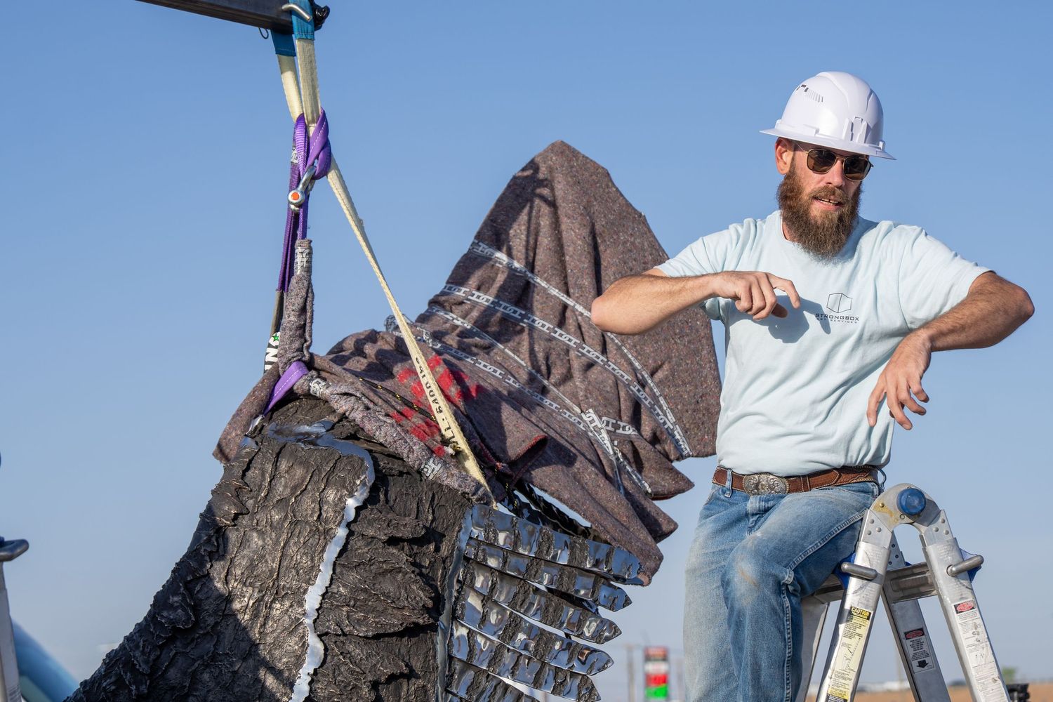 Bearded man in white hard hat and sunglasses posing beside a large dark sculpture outdoors on a clear day.
