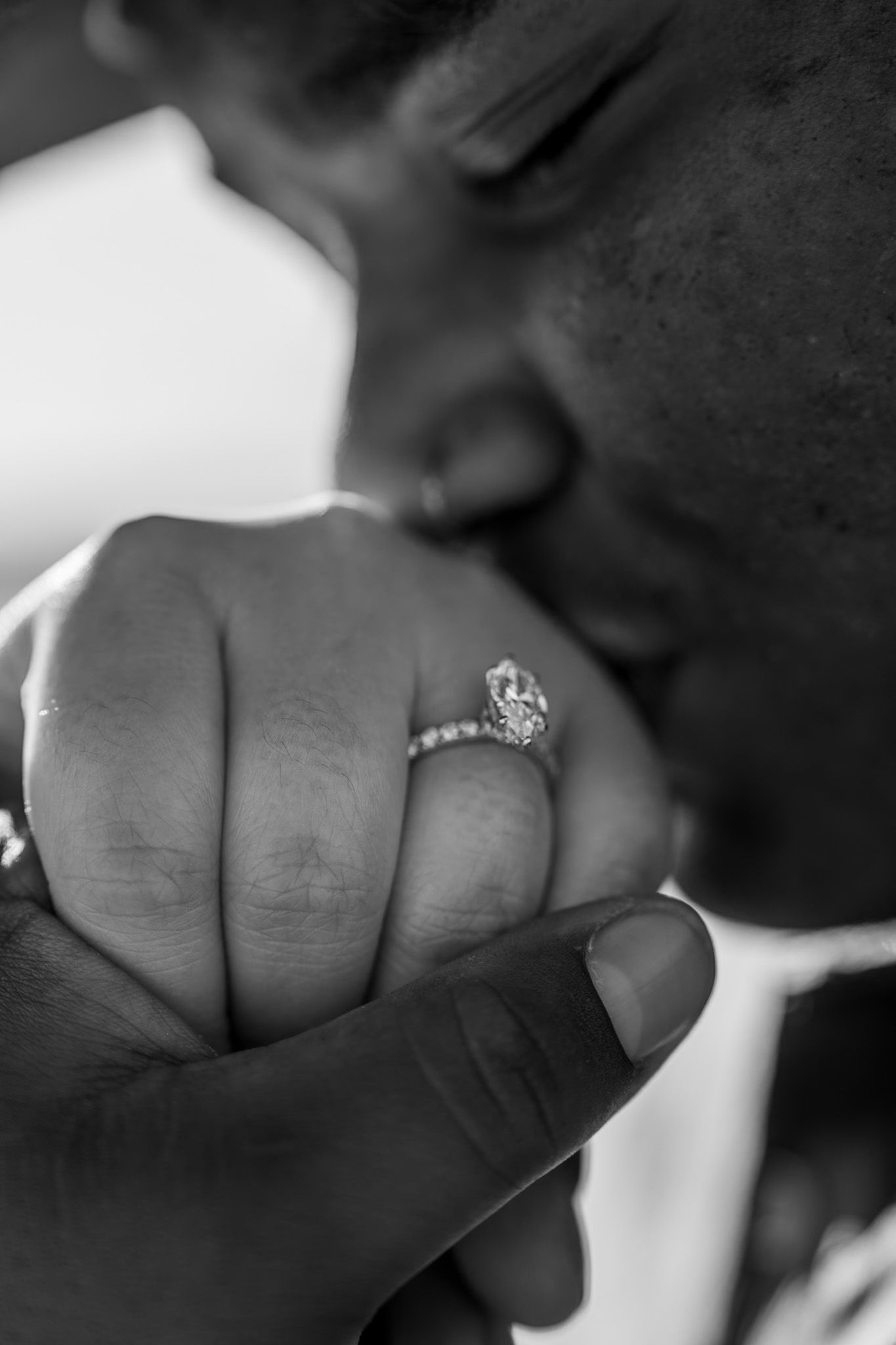 Black and white close-up of a person kissing a hand adorned with a diamond engagement ring.