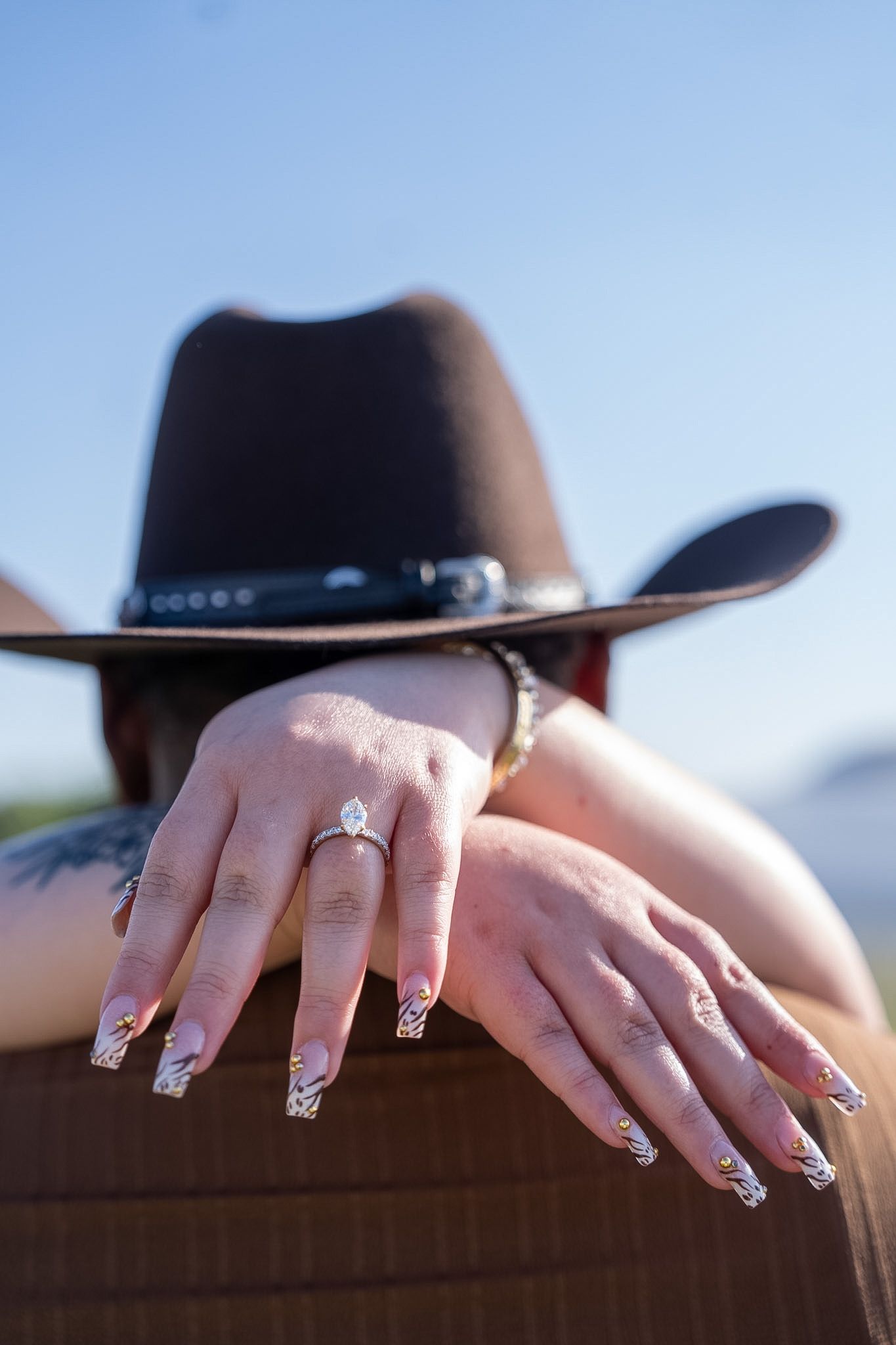 Close-up of hands with decorative nails and diamond ring resting on a surface with cowboy hat.