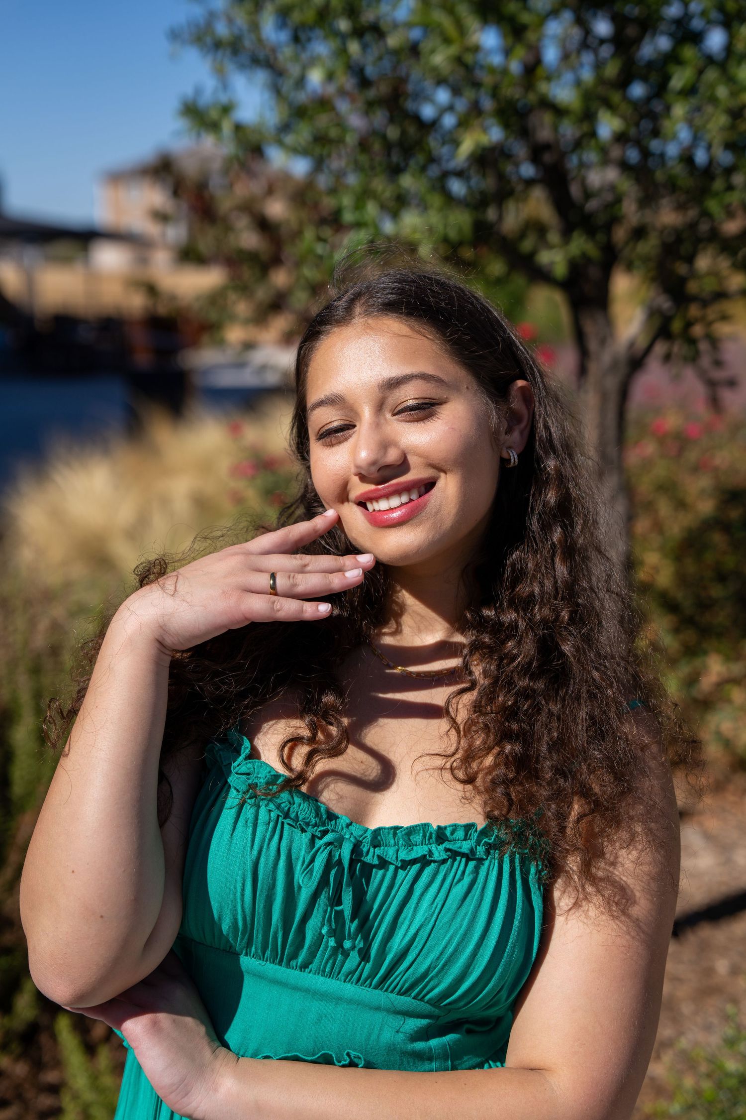 Smiling young woman in teal ruffled strapless dress poses outdoors with hand near face in sunny garden.