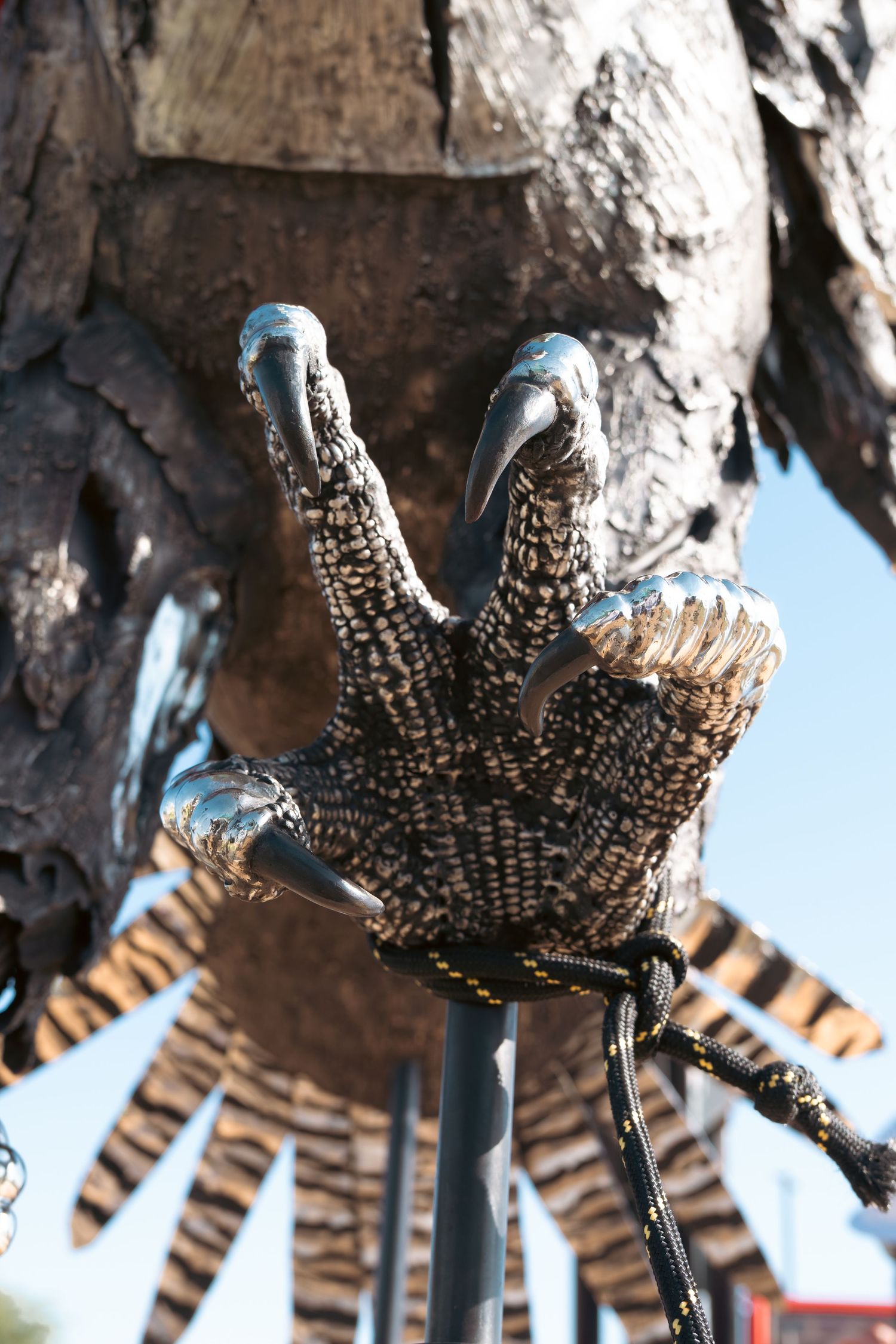 Close-up of a detailed metal bird talon sculpture gripping a pole, with textured feather patterns and shiny chrome claws.