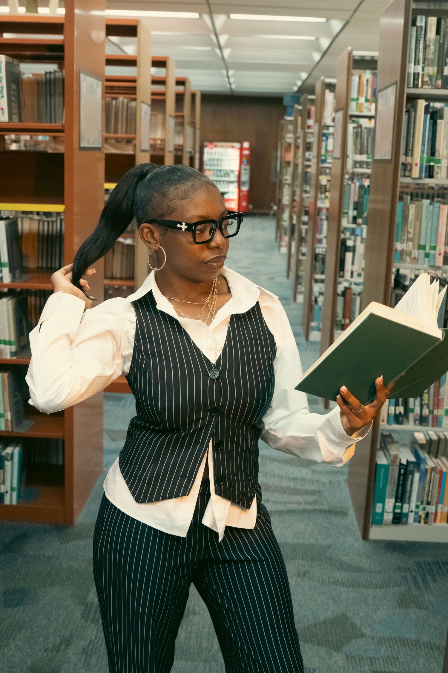 A Black woman in pinstripe vest and glasses reads a book while standing between library shelves.