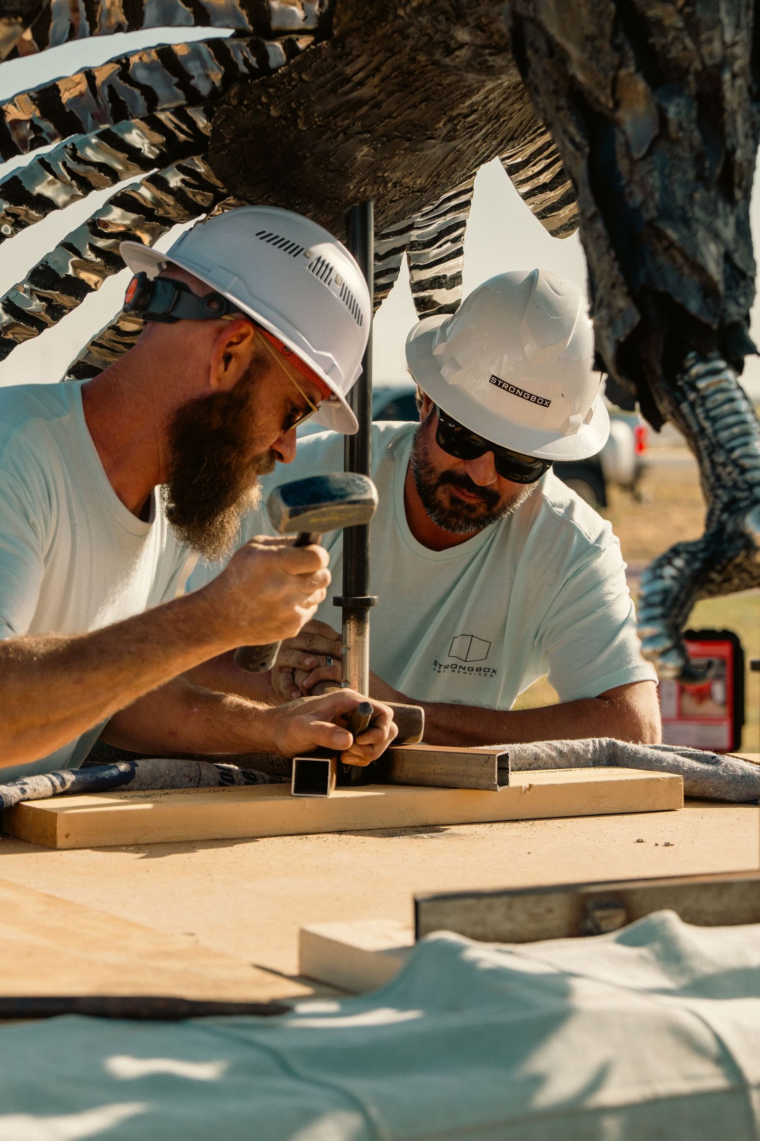 Two workers in hard hats collaborating on woodworking at outdoor construction site near metal sculpture.