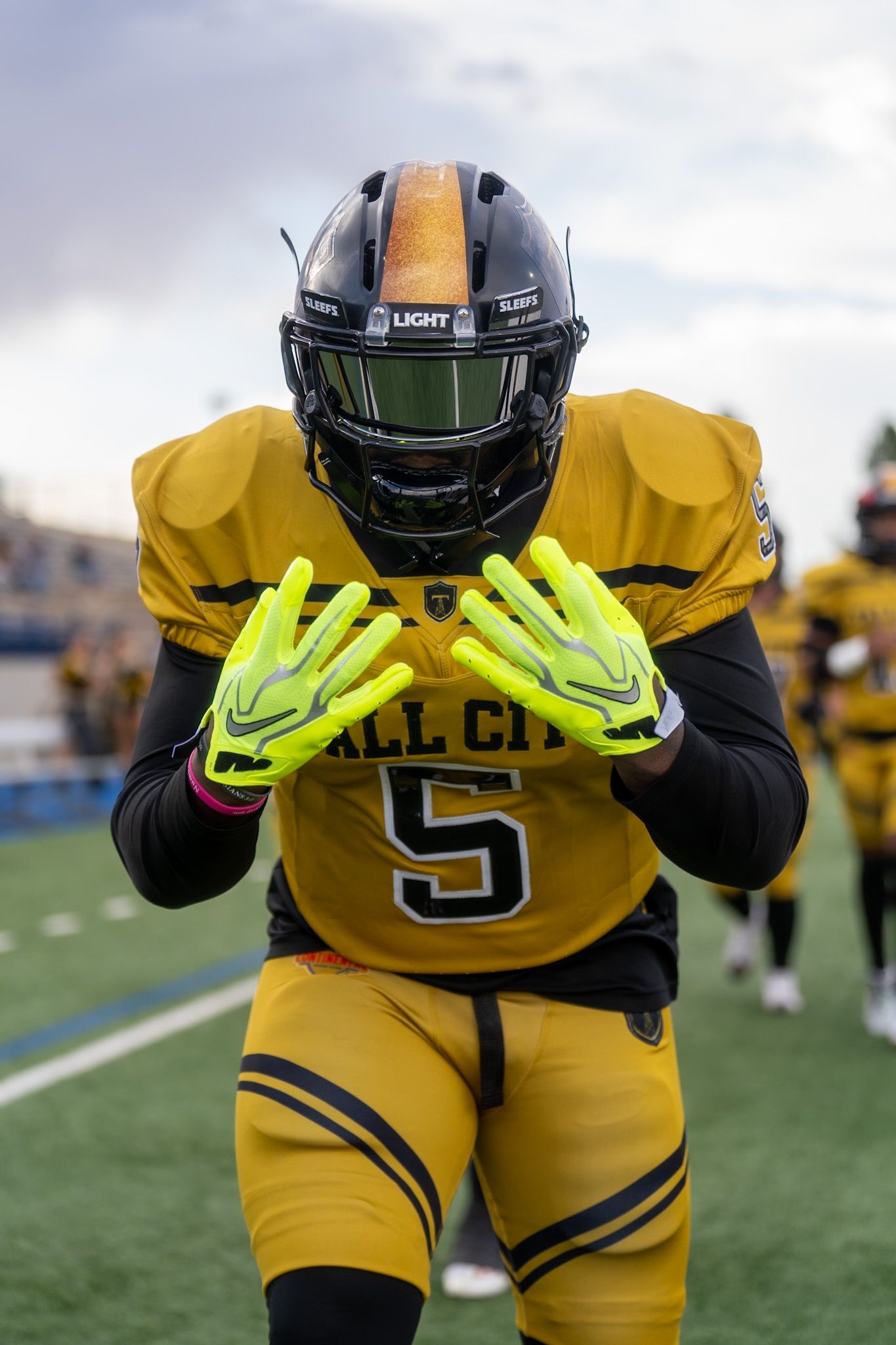 A football player in gold Tall City number 5 uniform holds up gloved hands on the field.