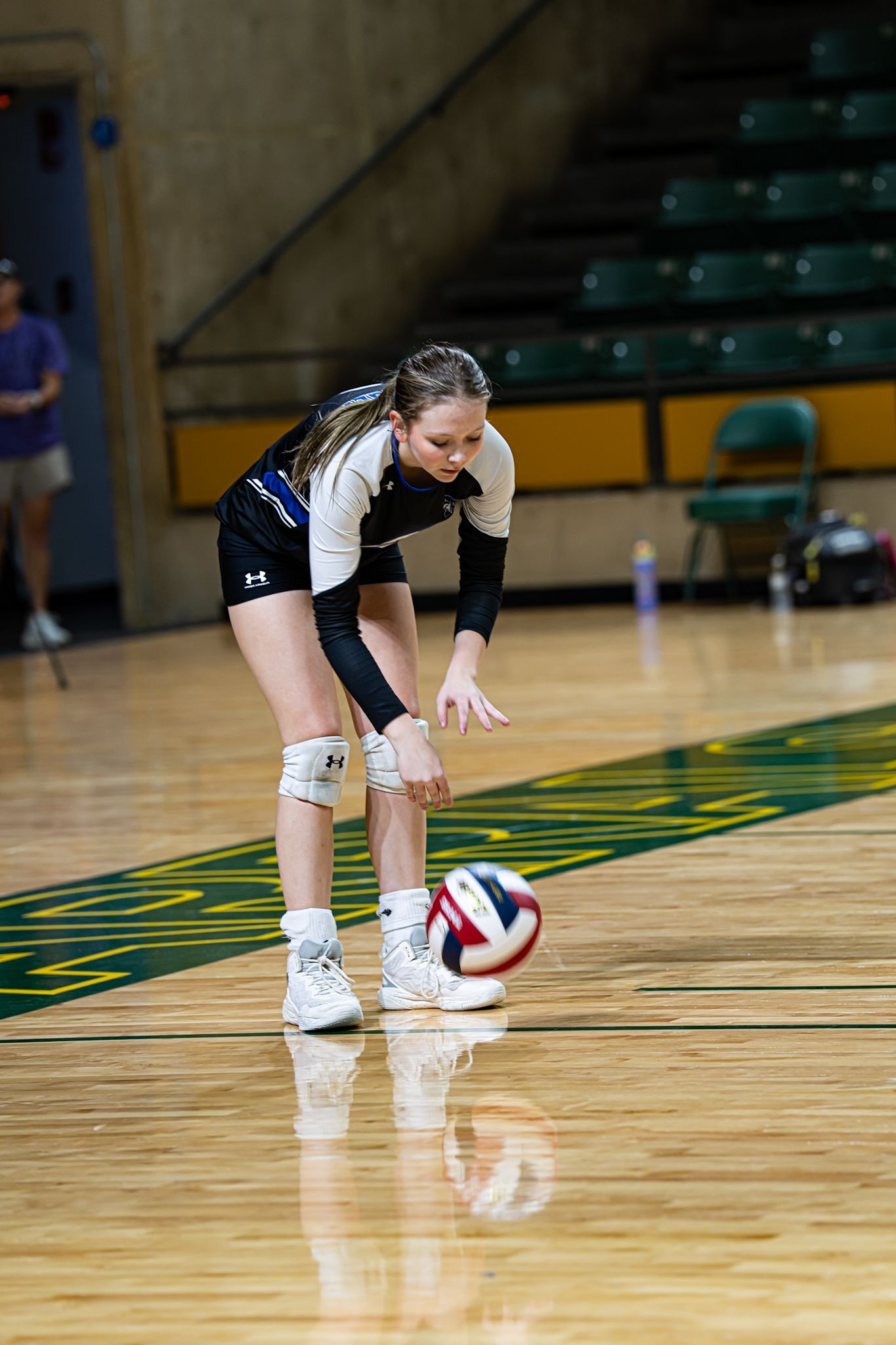 Volleyball player performs a dig, reaching low to receive ball on hardwood court.