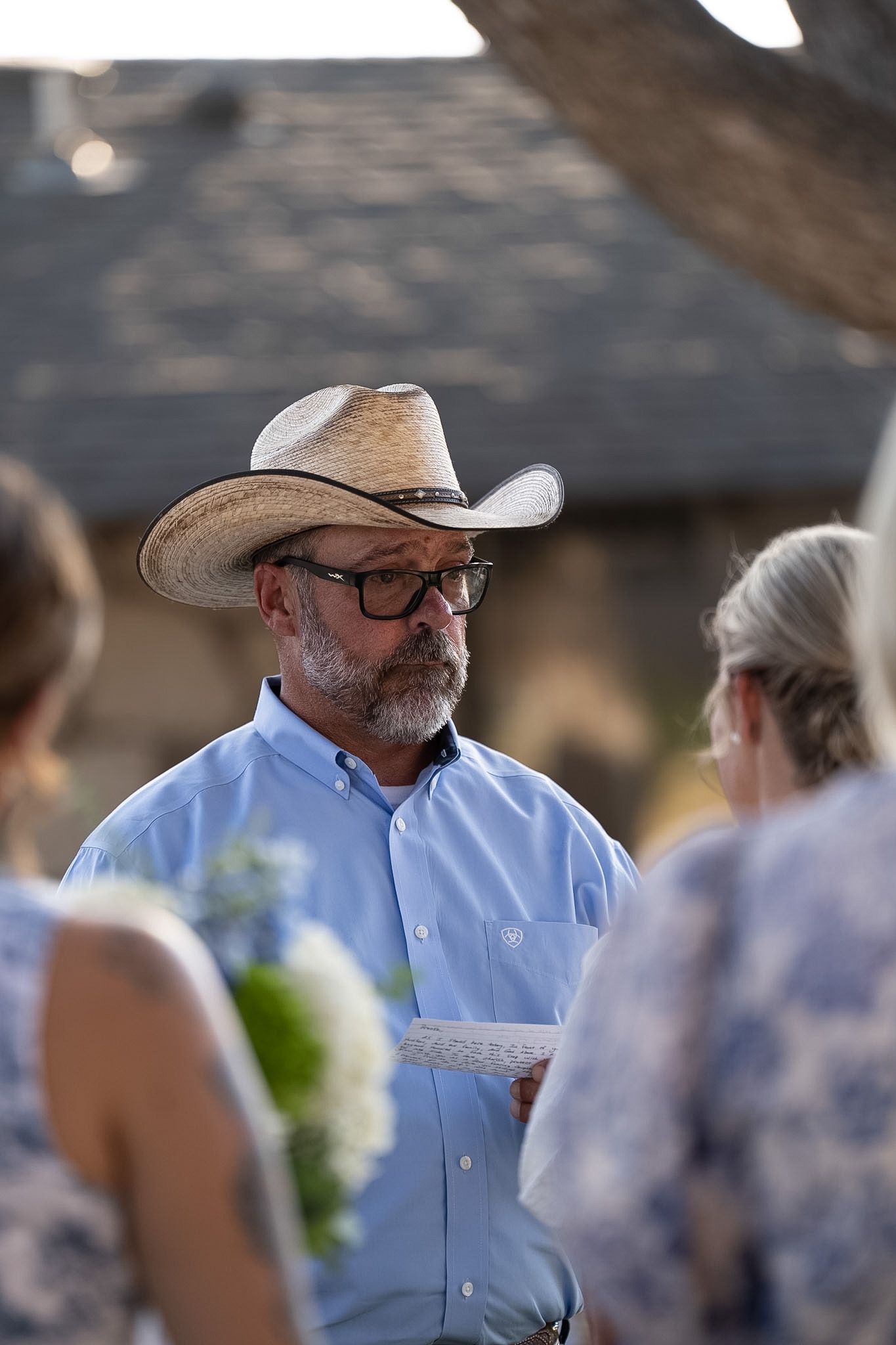Man in straw cowboy hat and glasses reads vows at outdoor wedding ceremony.