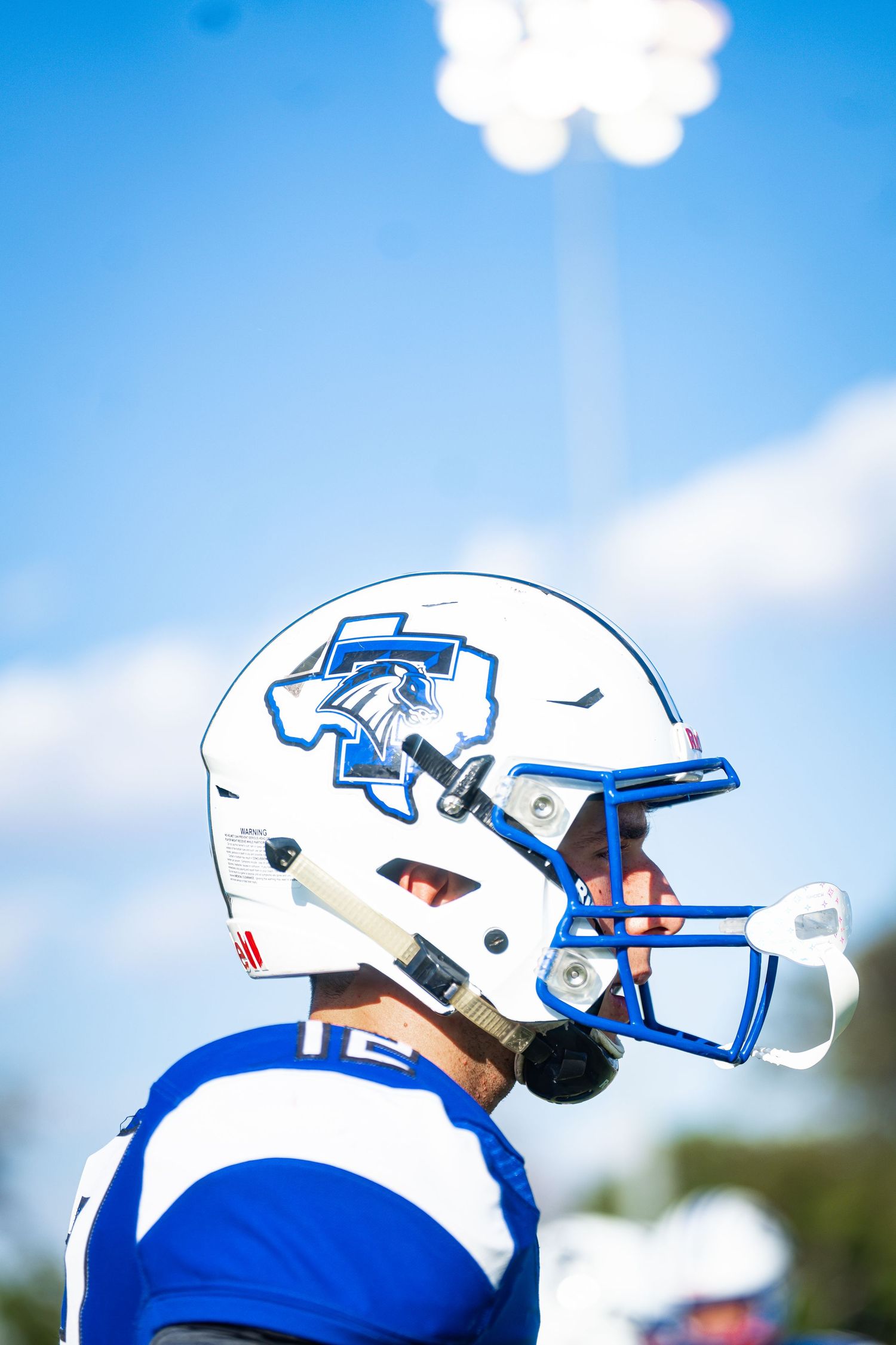 Football player wearing a white and blue helmet with a Texas logo and blue face mask under a bright sky.