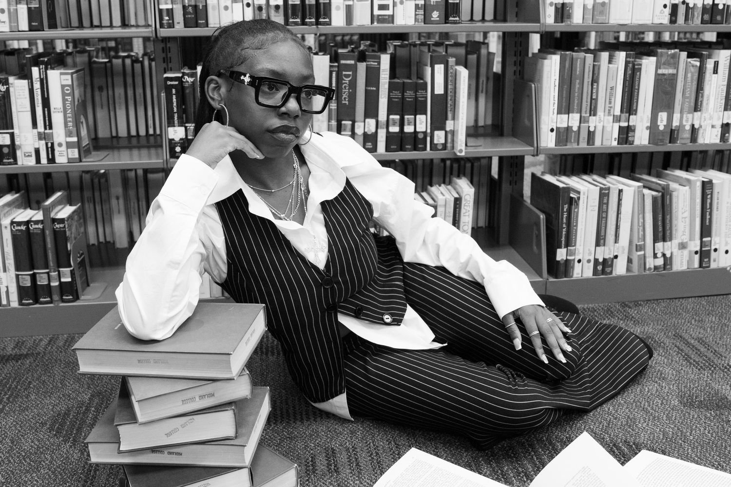 Black and white photo of stylish woman in pinstripe suit sitting on books on library floor.