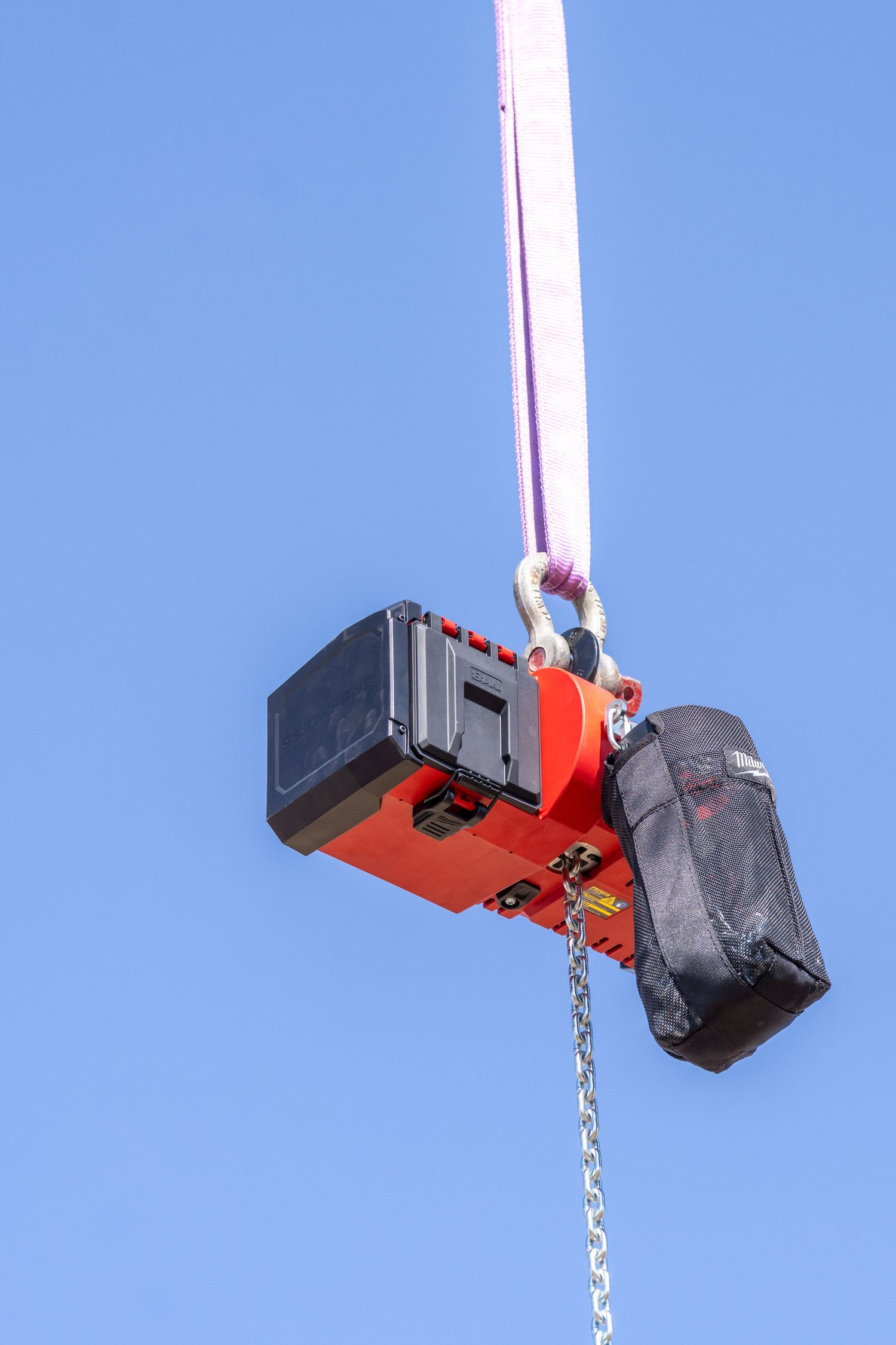 Red crane hook with black equipment case and chain hanging against clear blue sky.