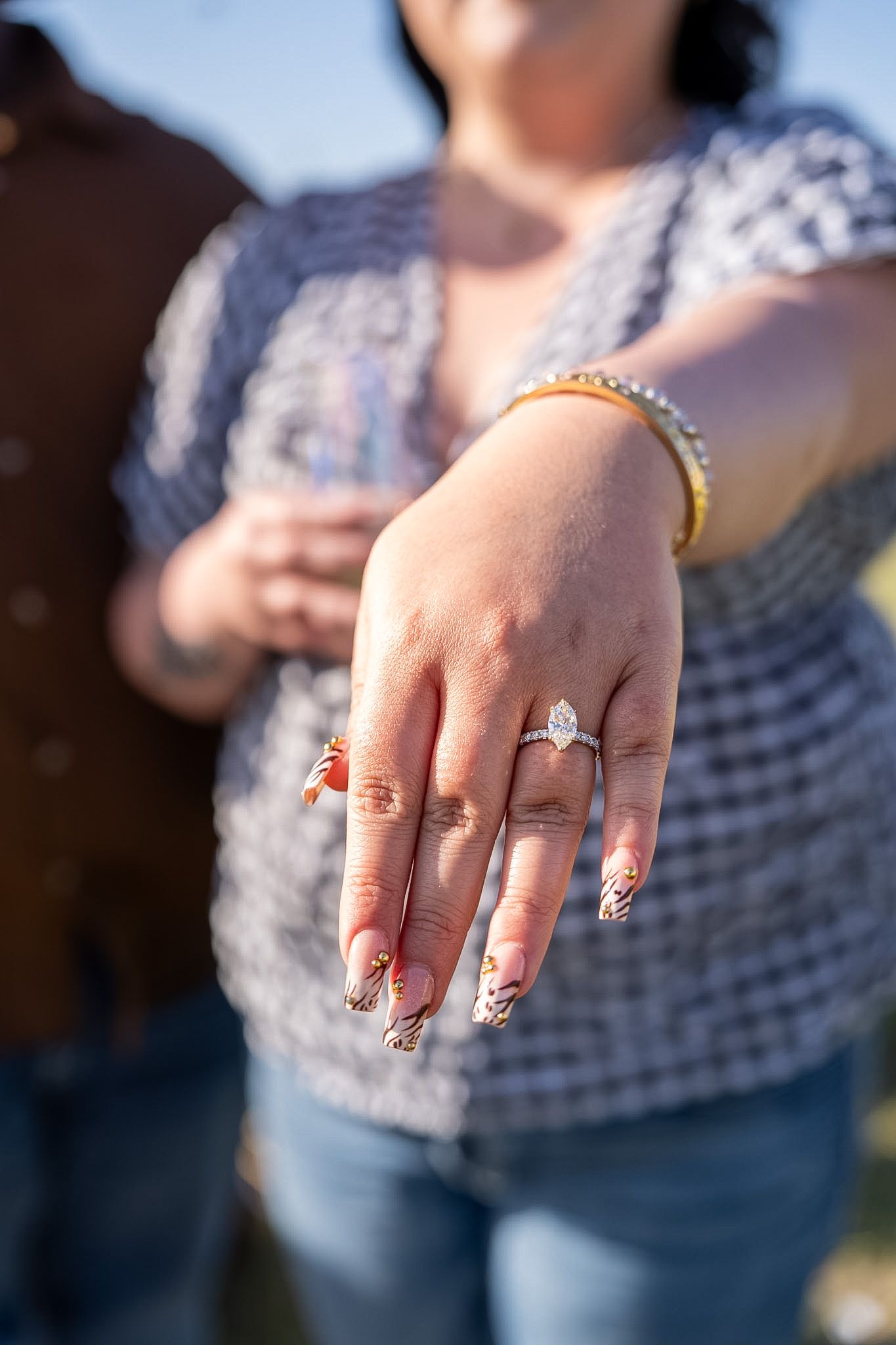 Woman showing off marquise diamond engagement ring with decorative nails and gold bracelet outdoors.