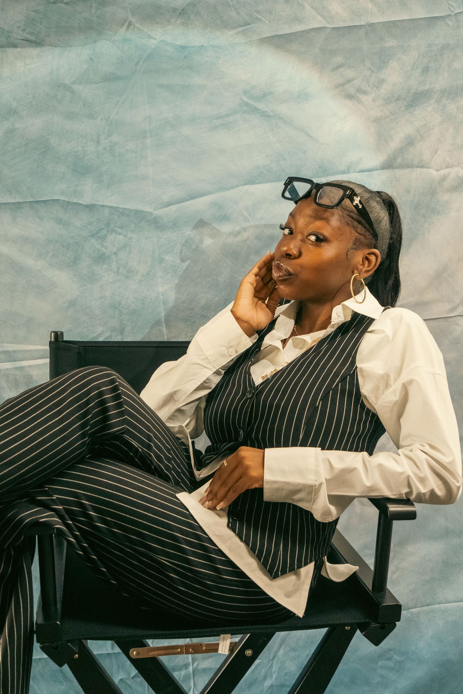 Stylish Black woman in pinstripe suit sits relaxed in director chair against blue crinkled backdrop.