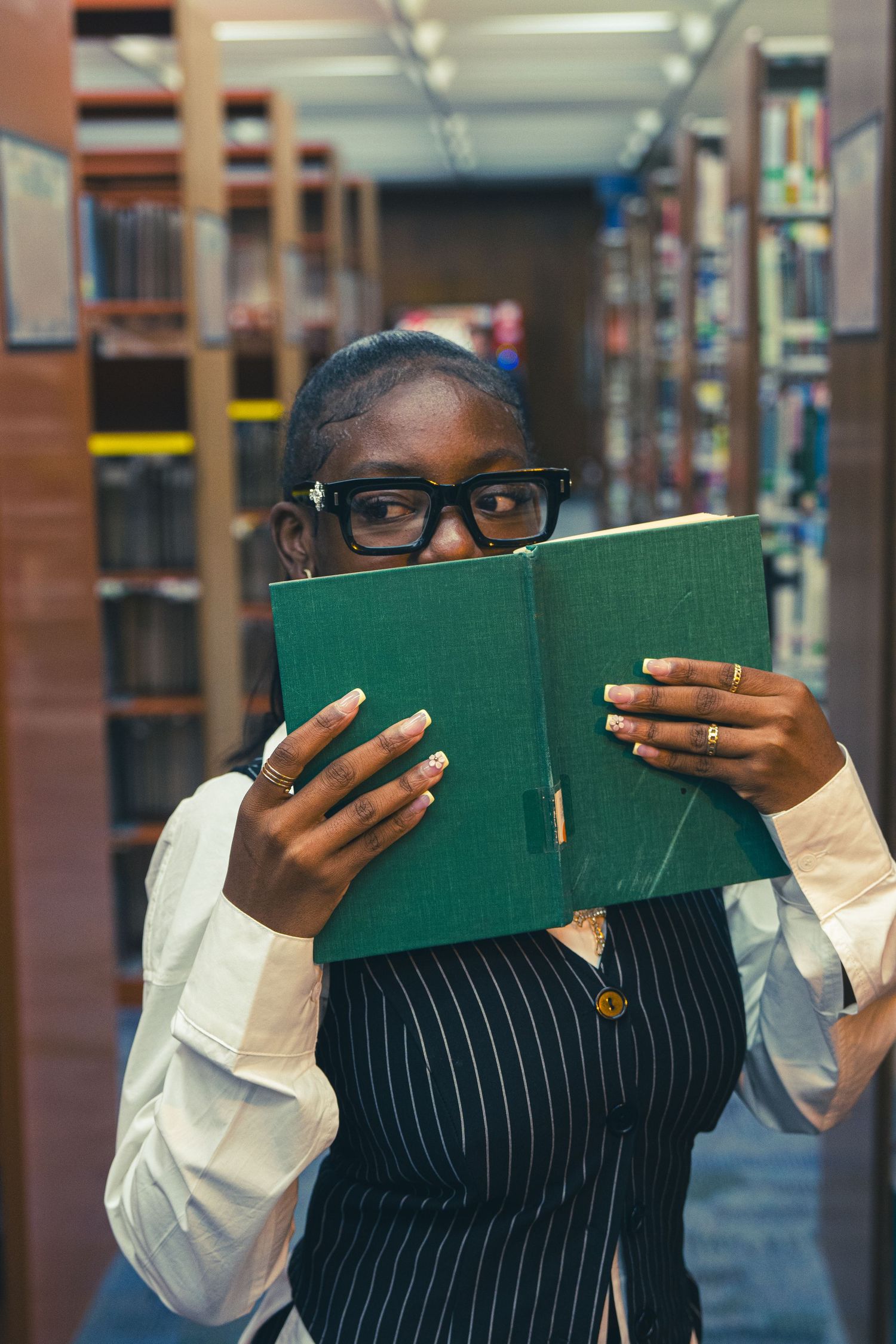 Woman in pinstripe vest and white shirt holds green book over face in colorful library aisle.
