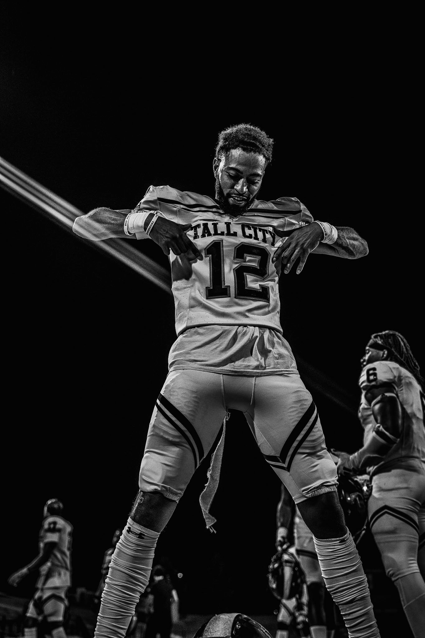 Football player #12 in Tall City jersey poses dramatically under spotlight in black and white.