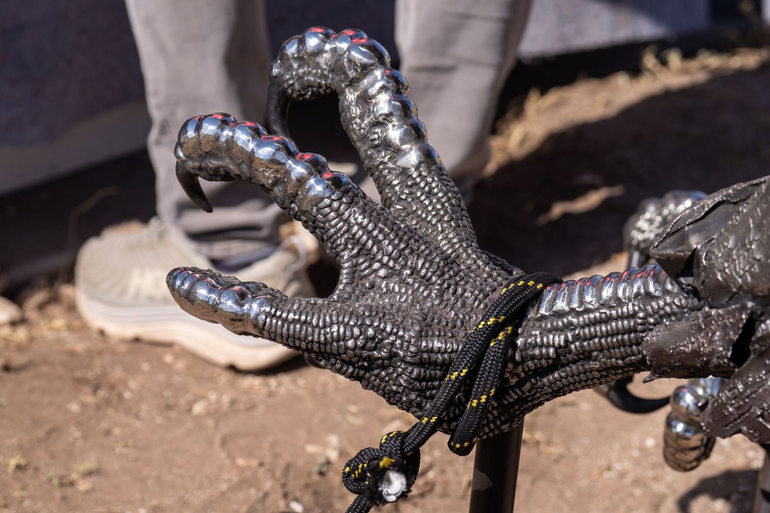 Close-up of a metallic decorative bird claw sculpture with textured scales, displayed outdoors on sandy ground.