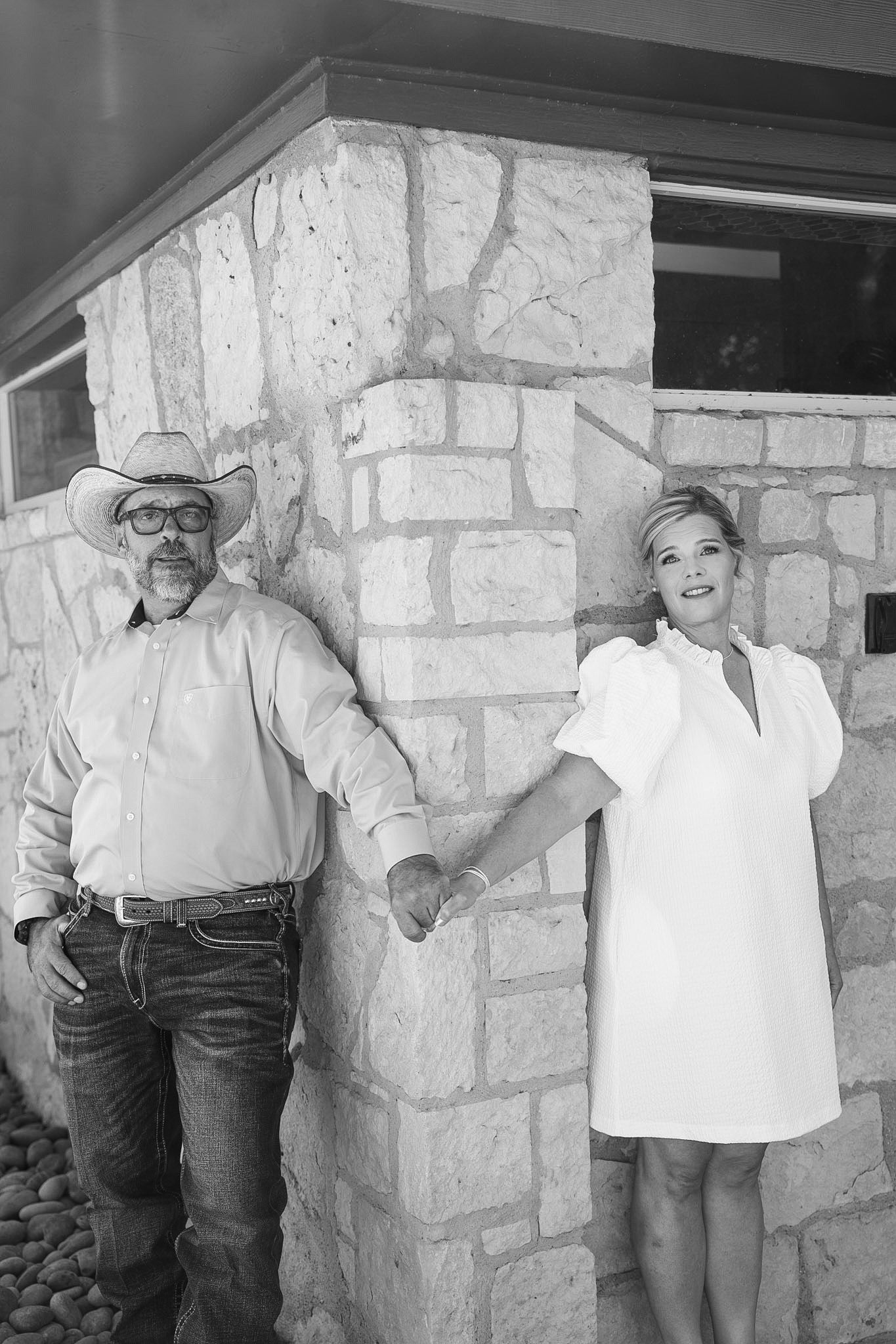 Black and white photo of couple holding hands by stone pillar, man in cowboy hat and woman in white dress.