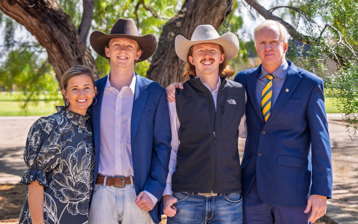 Four people smiling outdoors under a tree, dressed in smart casual attire including cowboy hats and blazers.