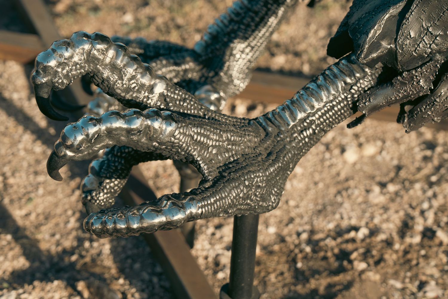 Close-up of a metallic dragon claw sculpture with detailed scales, displayed outdoors on a sandy surface.