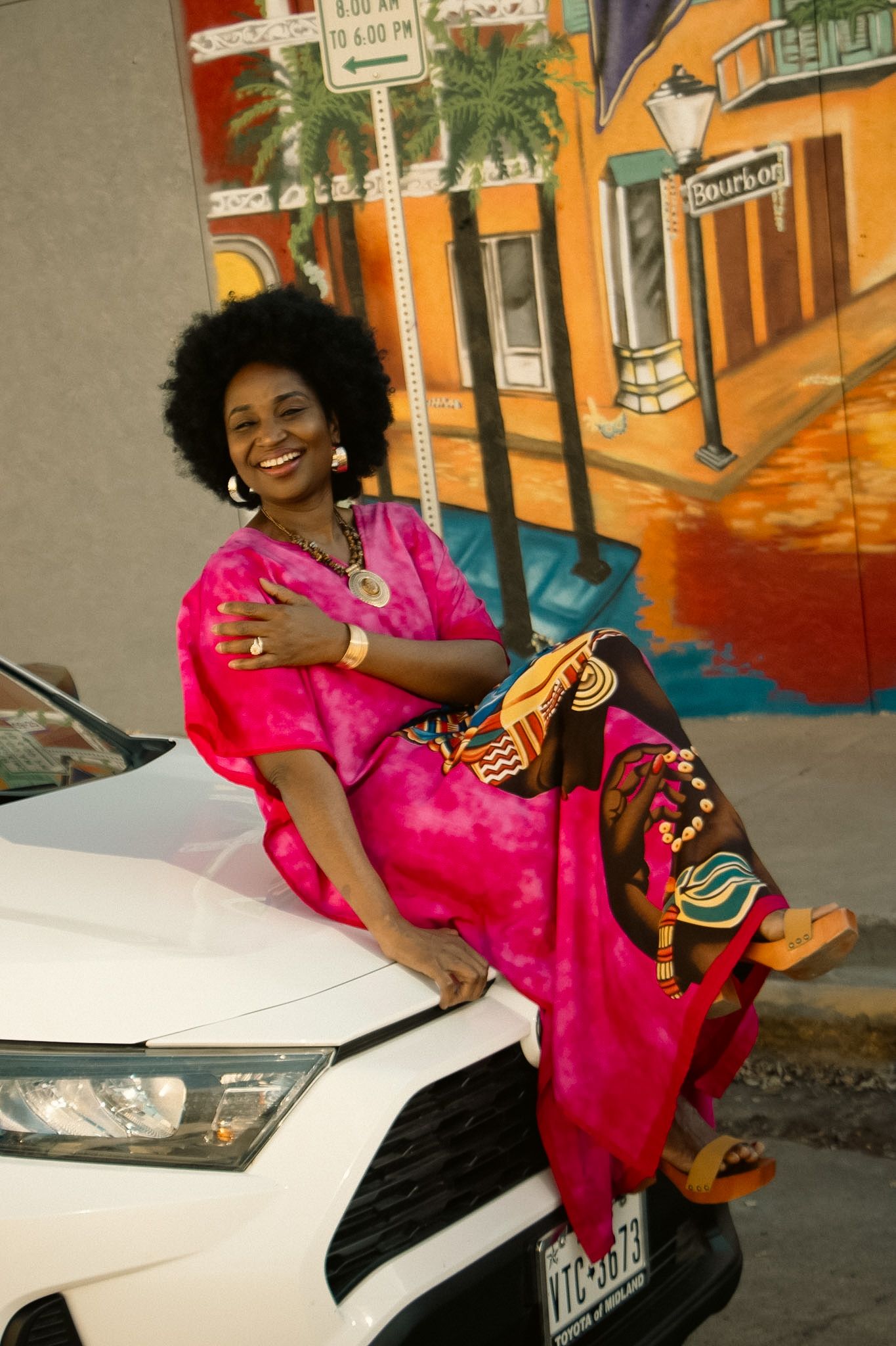 Woman in pink African dress sits on white car before Bourbon Street mural.