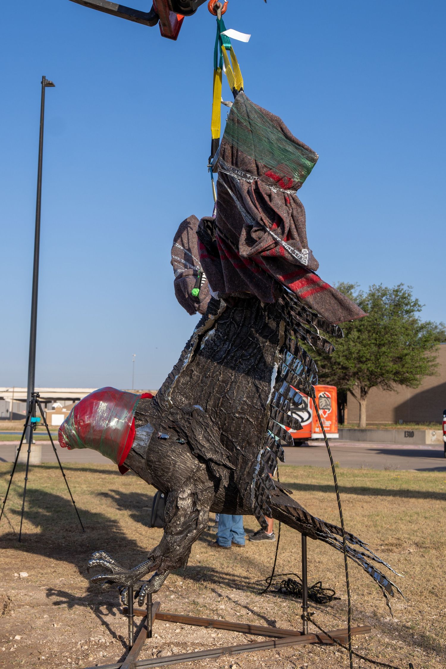 A large dinosaur sculpture being hoisted by yellow straps on a metal frame outdoors under a clear blue sky.