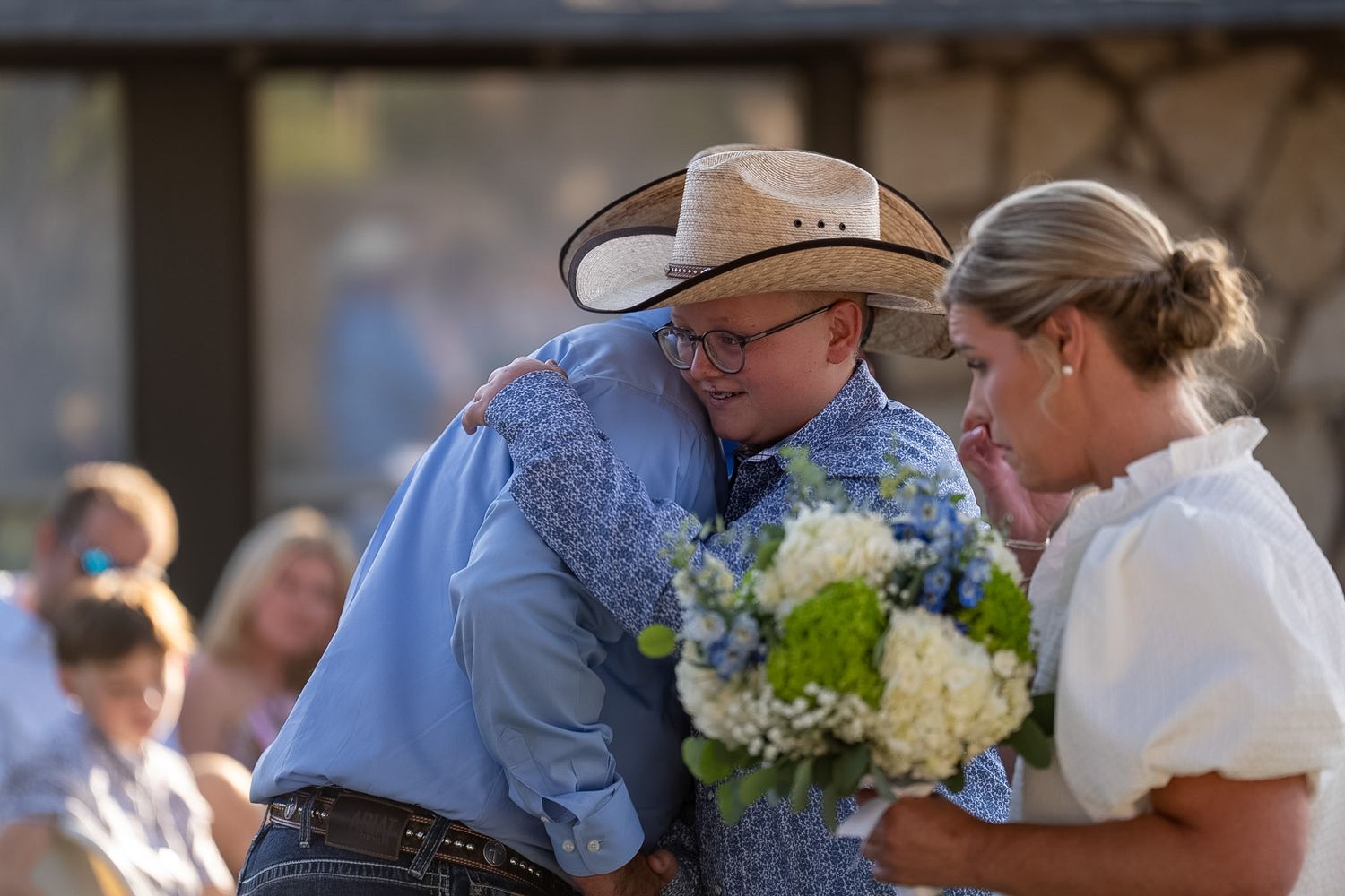 Young man in cowboy hat and blue shirt hugs woman holding white and blue bouquet at outdoor wedding.