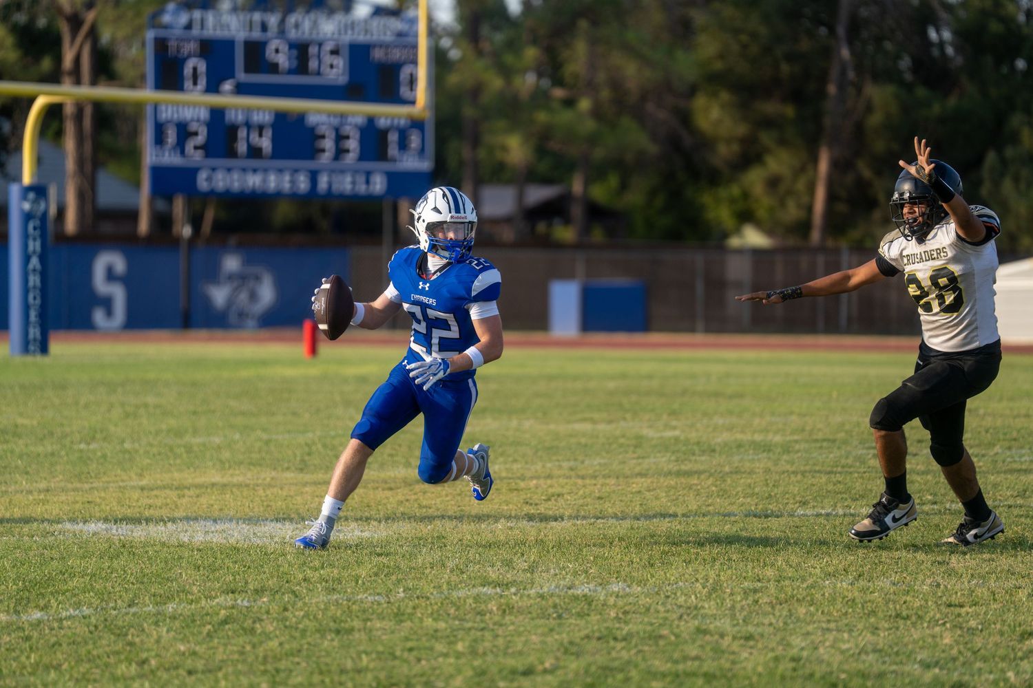 Youth football player in blue uniform runs with ball as defender in black attempts to block on green field.