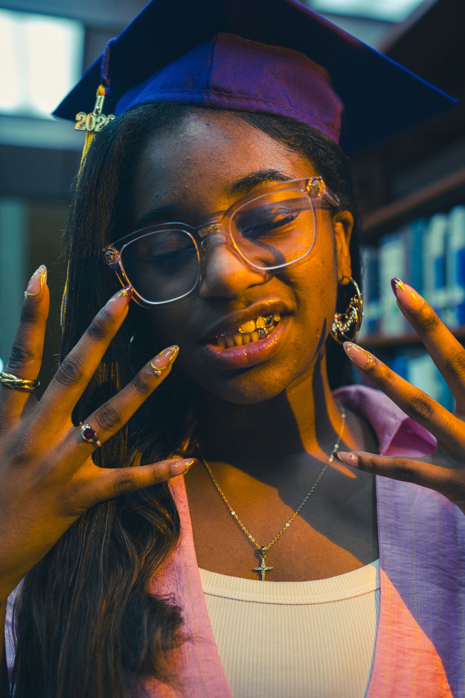 A graduating Black woman wearing a purple cap and gold grills shows her nails in a library setting.