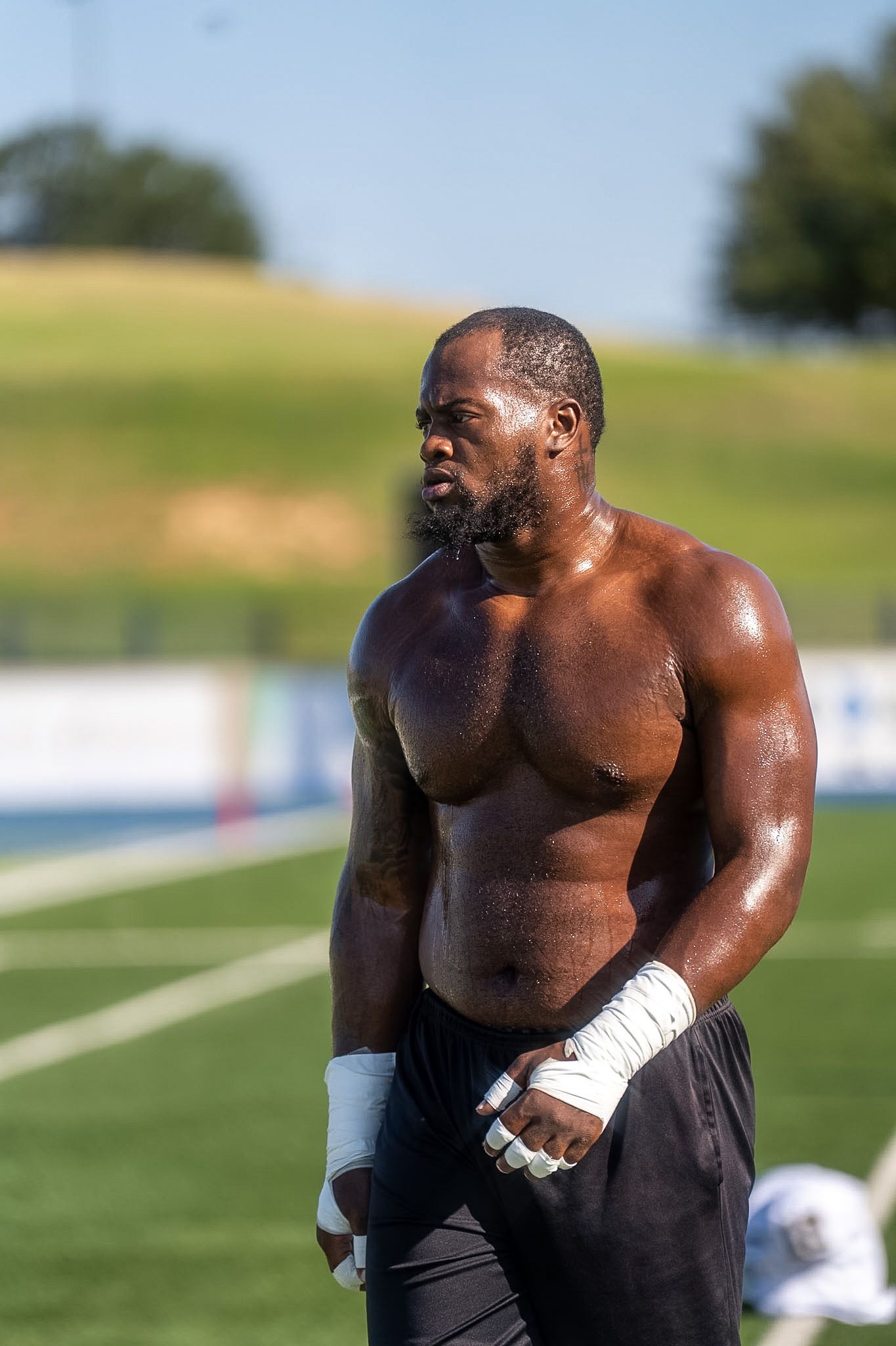 Muscular shirtless football player with wrapped wrists standing on green field after practice.