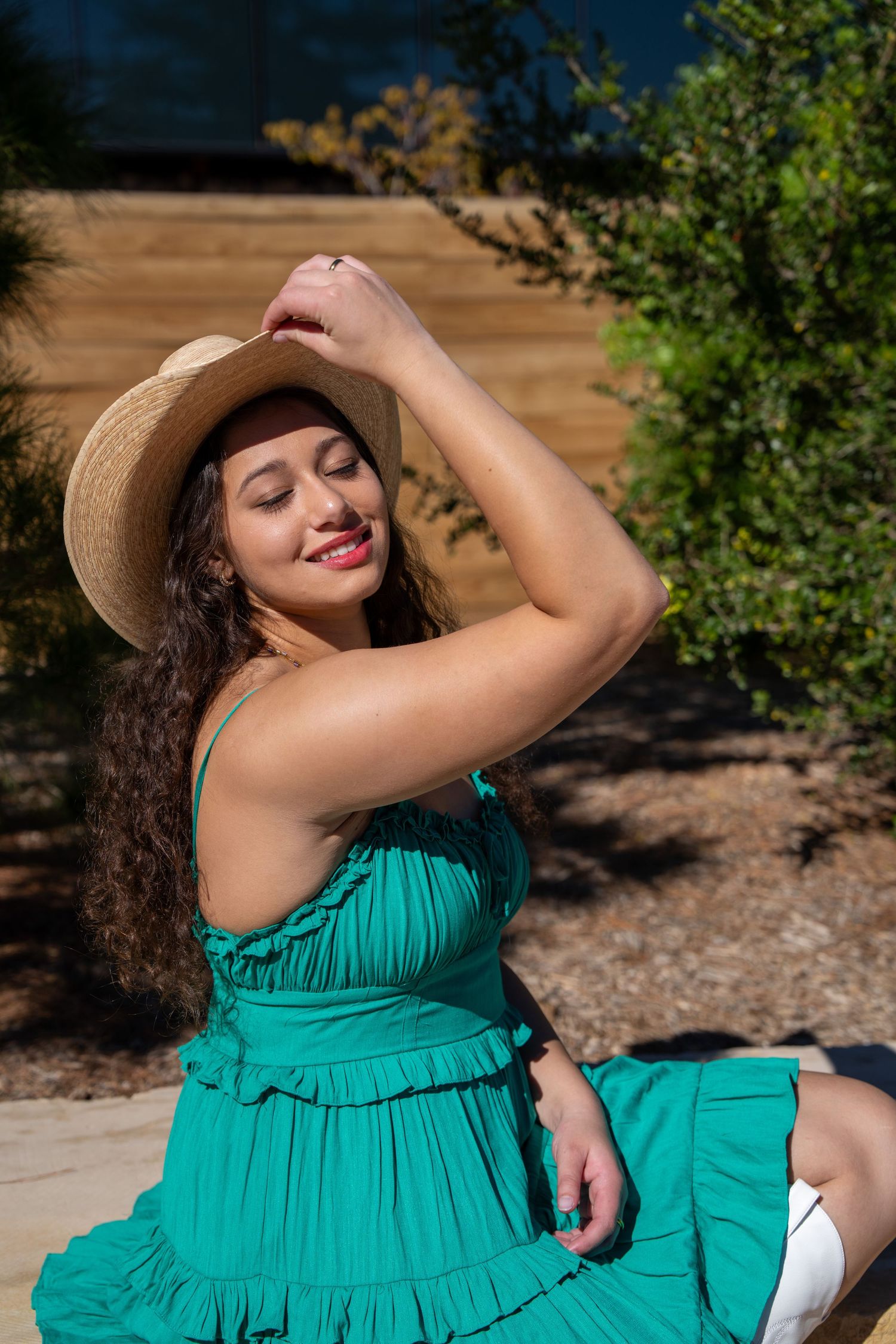 Young woman in green tiered sundress and straw hat sits outdoors smiling with eyes closed in sunlight.