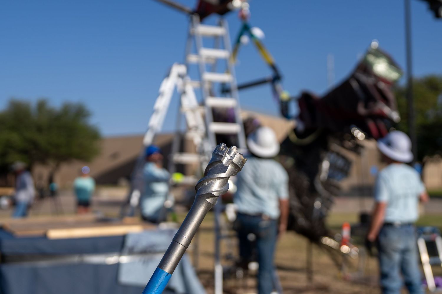 Workers install solar panels on a sunny day with a drill bit in focus and a ladder in the background.