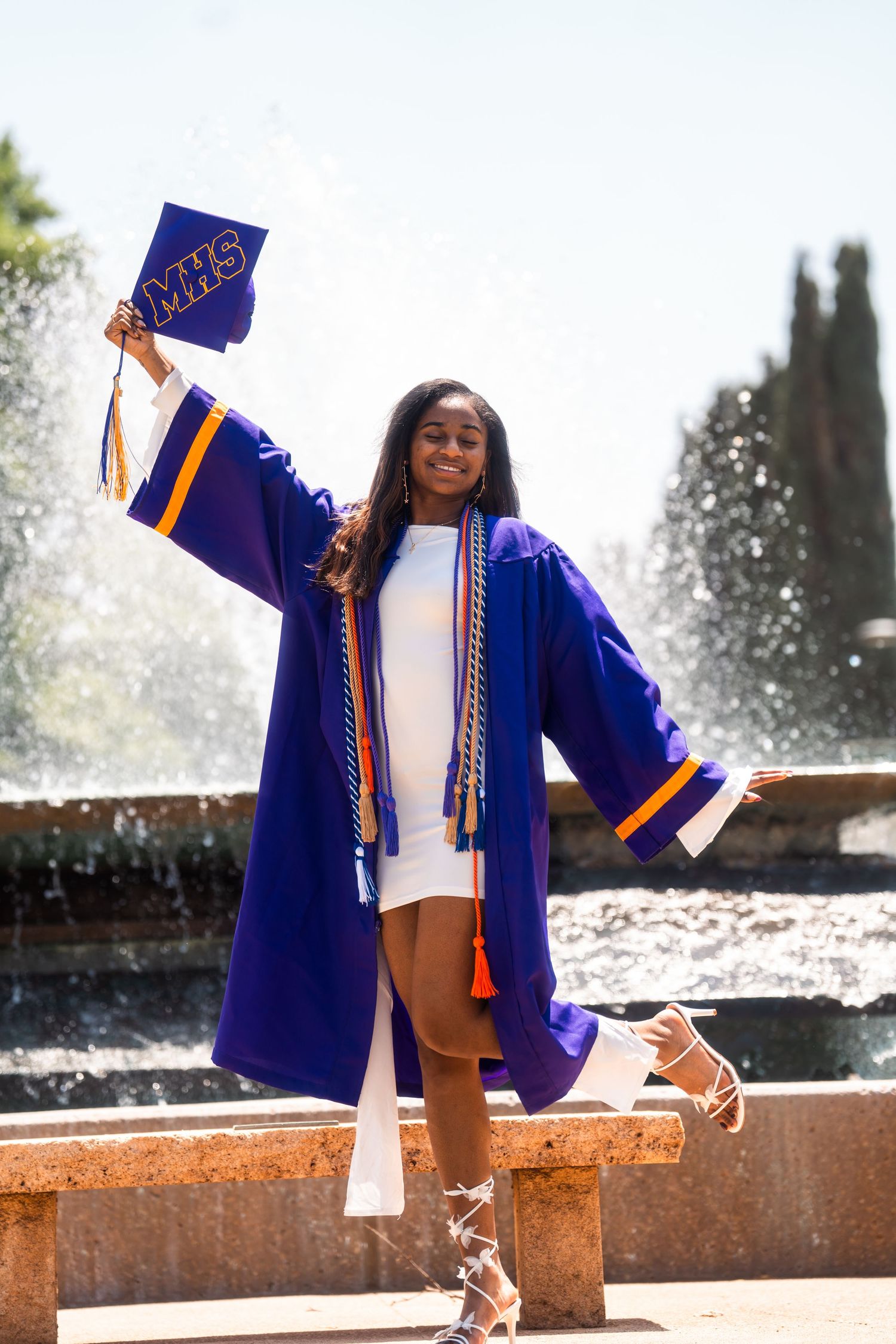 A smiling graduate in a blue cap and gown holds her MHS cap while posing in front of a fountain.