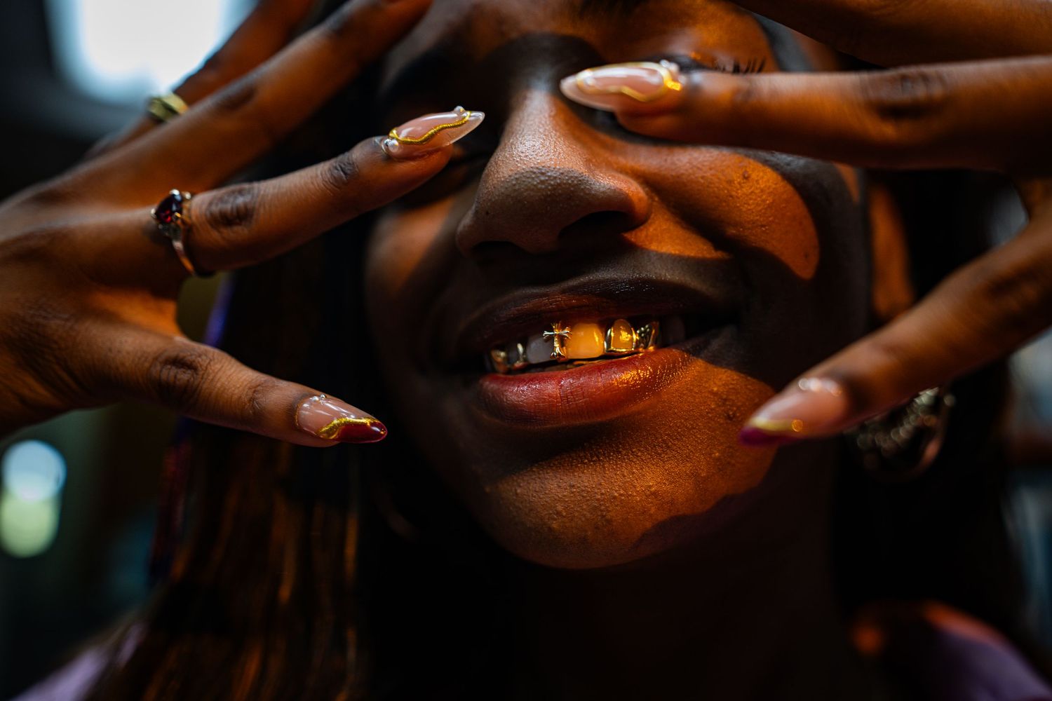 Close-up of a smiling person with gold grillz, long decorated nails framing their face, wearing multiple rings.