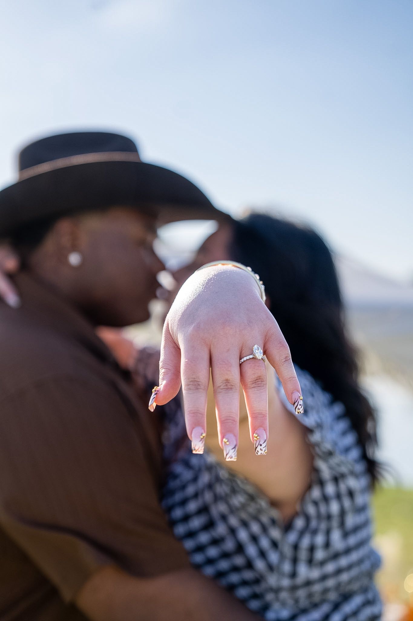 Couple kissing outdoors while woman displays diamond engagement ring toward camera.