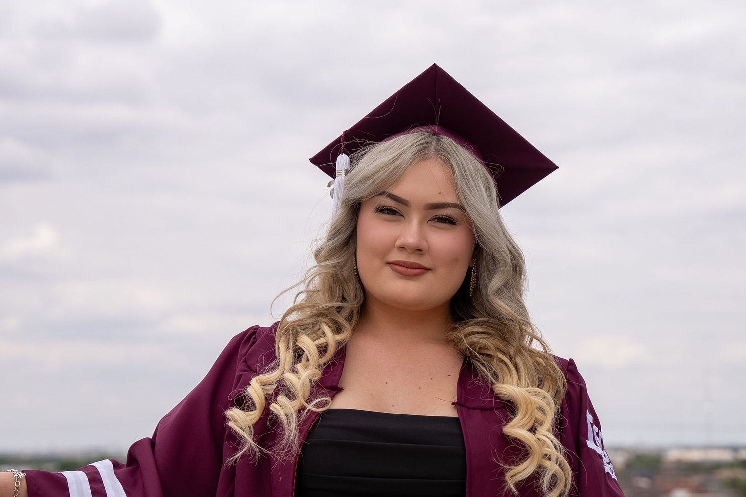 Young woman in maroon graduation cap and gown smiling outdoors on a cloudy day.