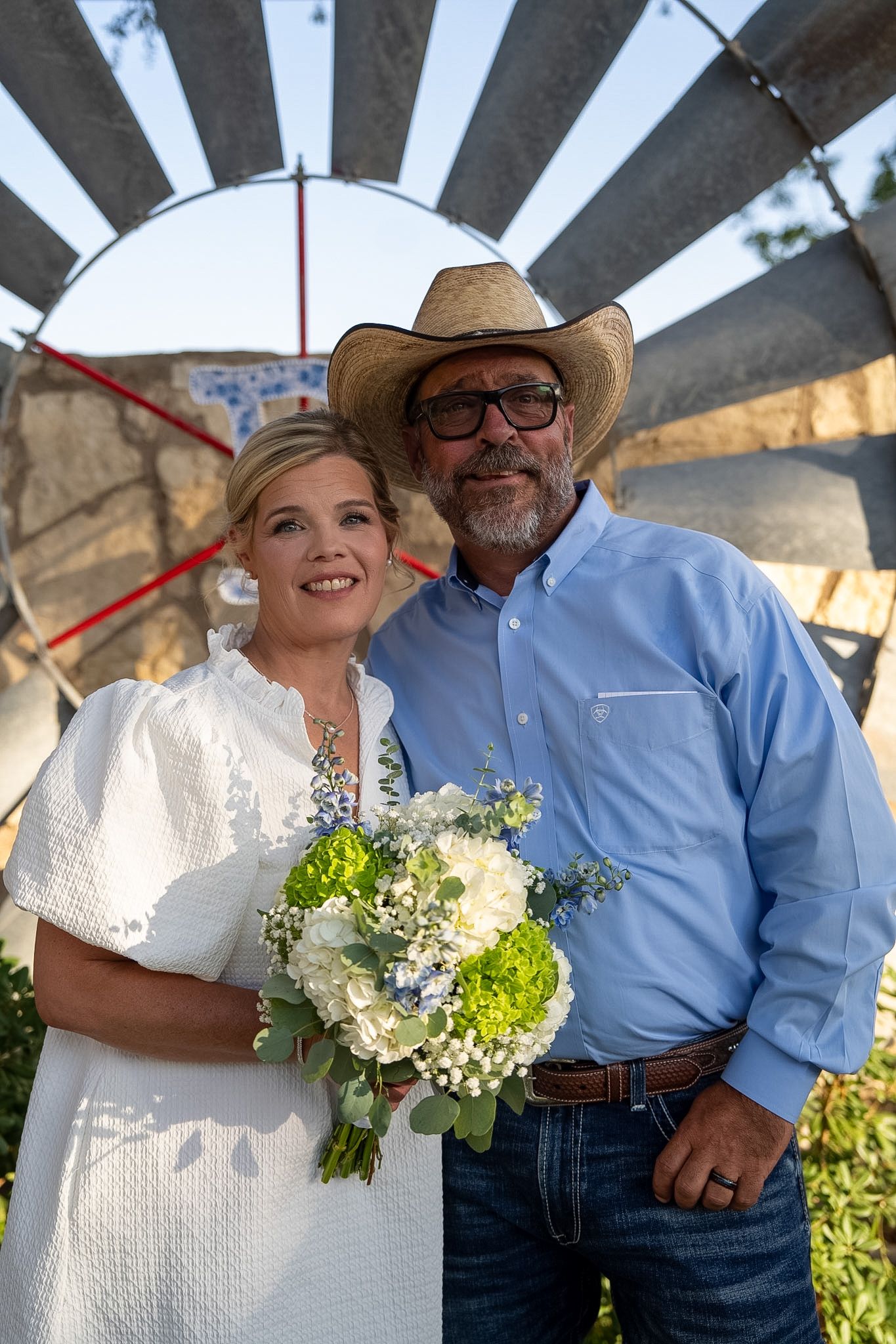 A couple poses outdoors, bride holding green and white bouquet, groom in cowboy hat and blue shirt, windmill behind them.