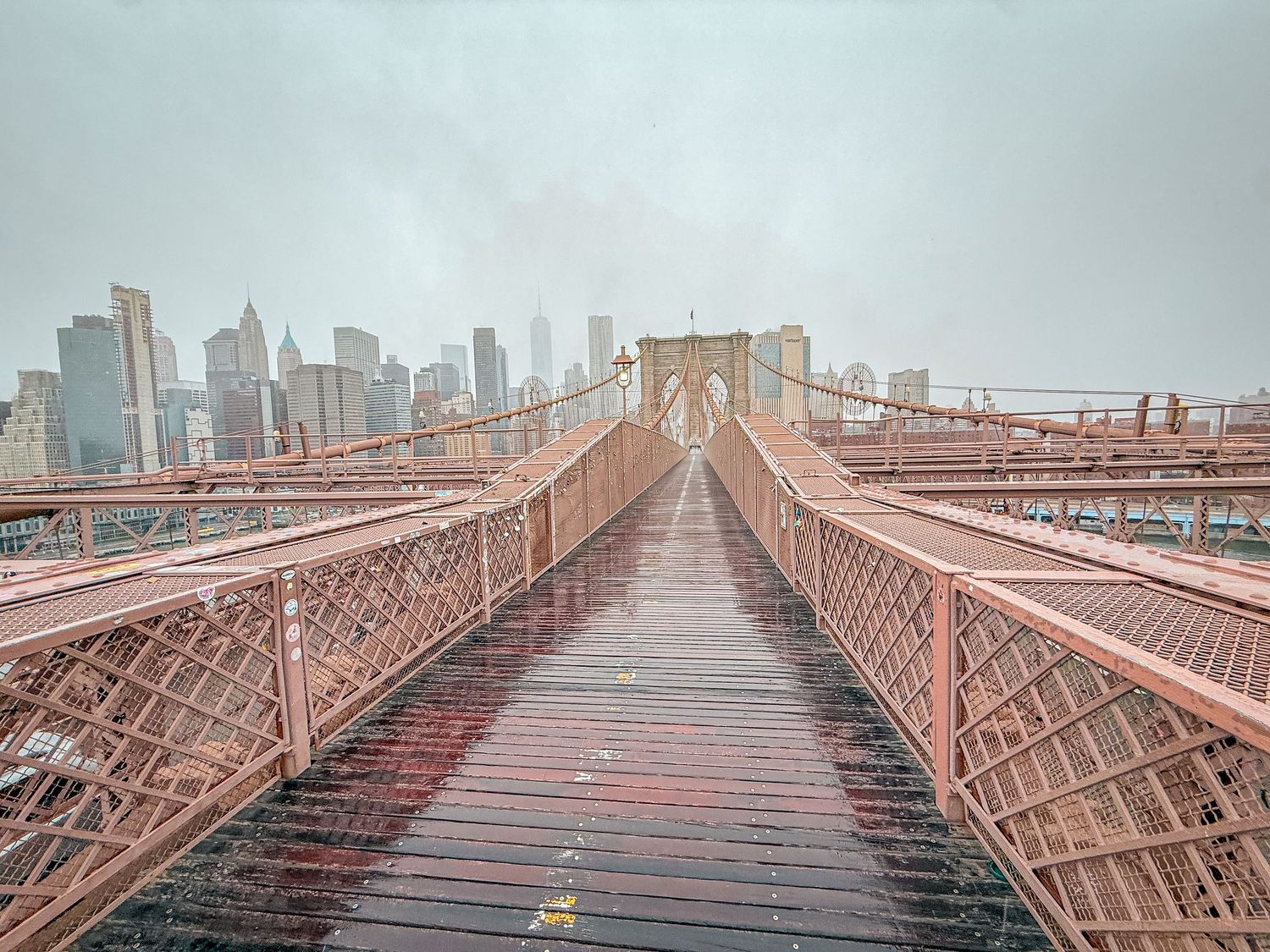 Rainy day view on the Brooklyn Bridge walkway with wet wooden planks and Manhattan skyline in the misty background.