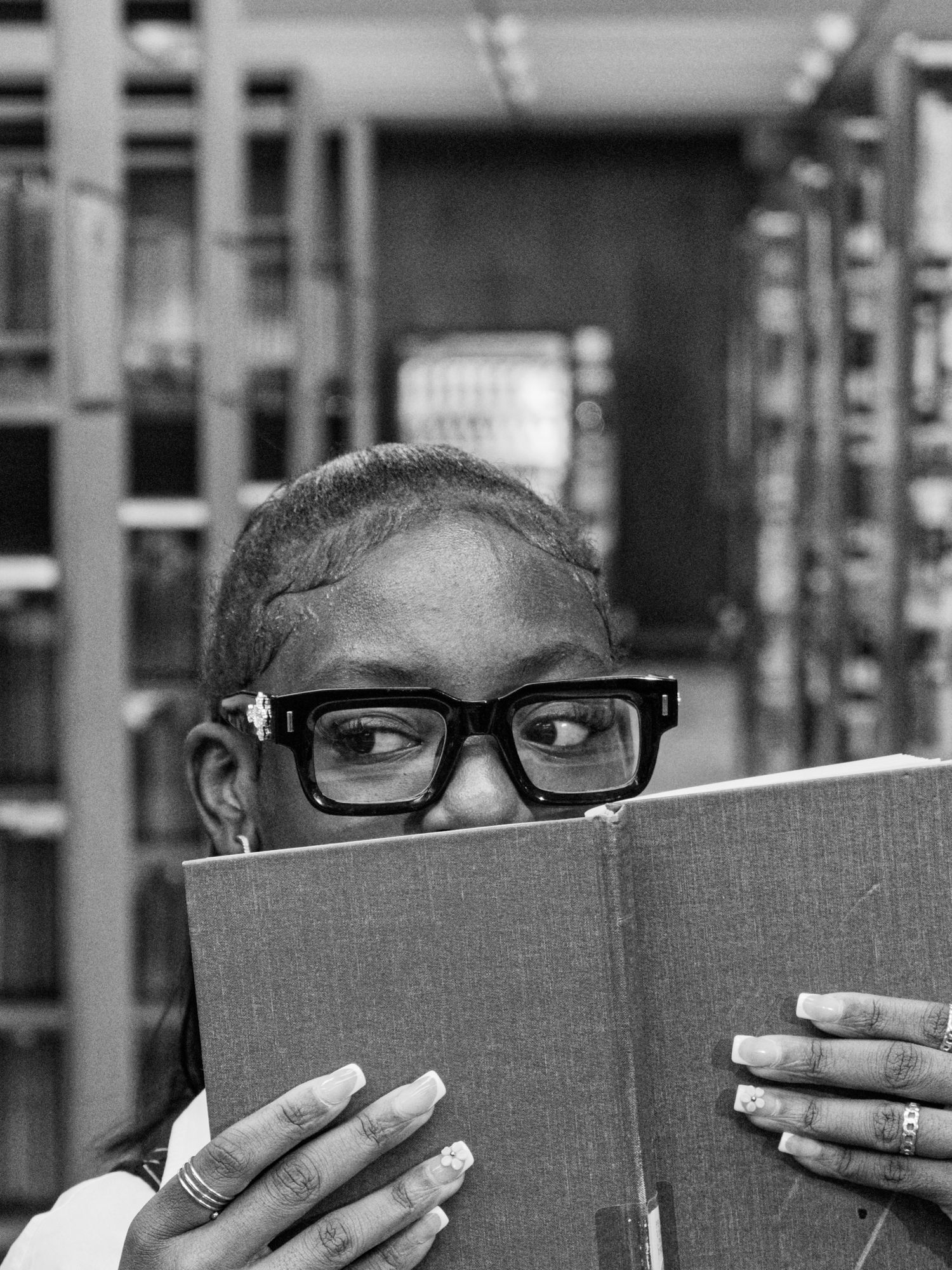 A young woman with glasses reads a book in a library, her face partially hidden behind the open pages.