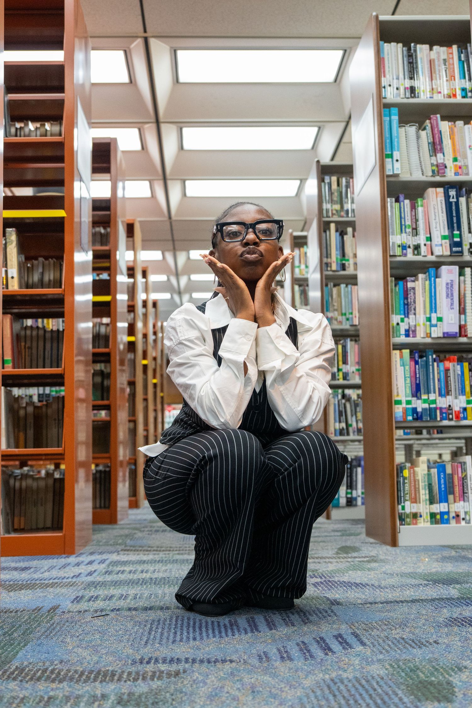 A stylish woman in glasses and pinstripe pants crouches between library bookshelves, posing with hands framing her face.