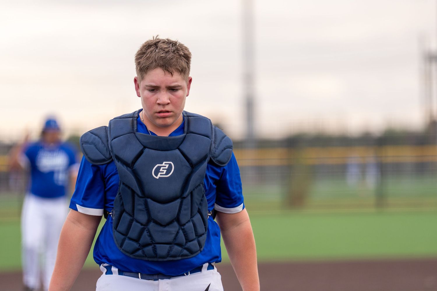 Young baseball catcher in blue uniform with chest protector on field.