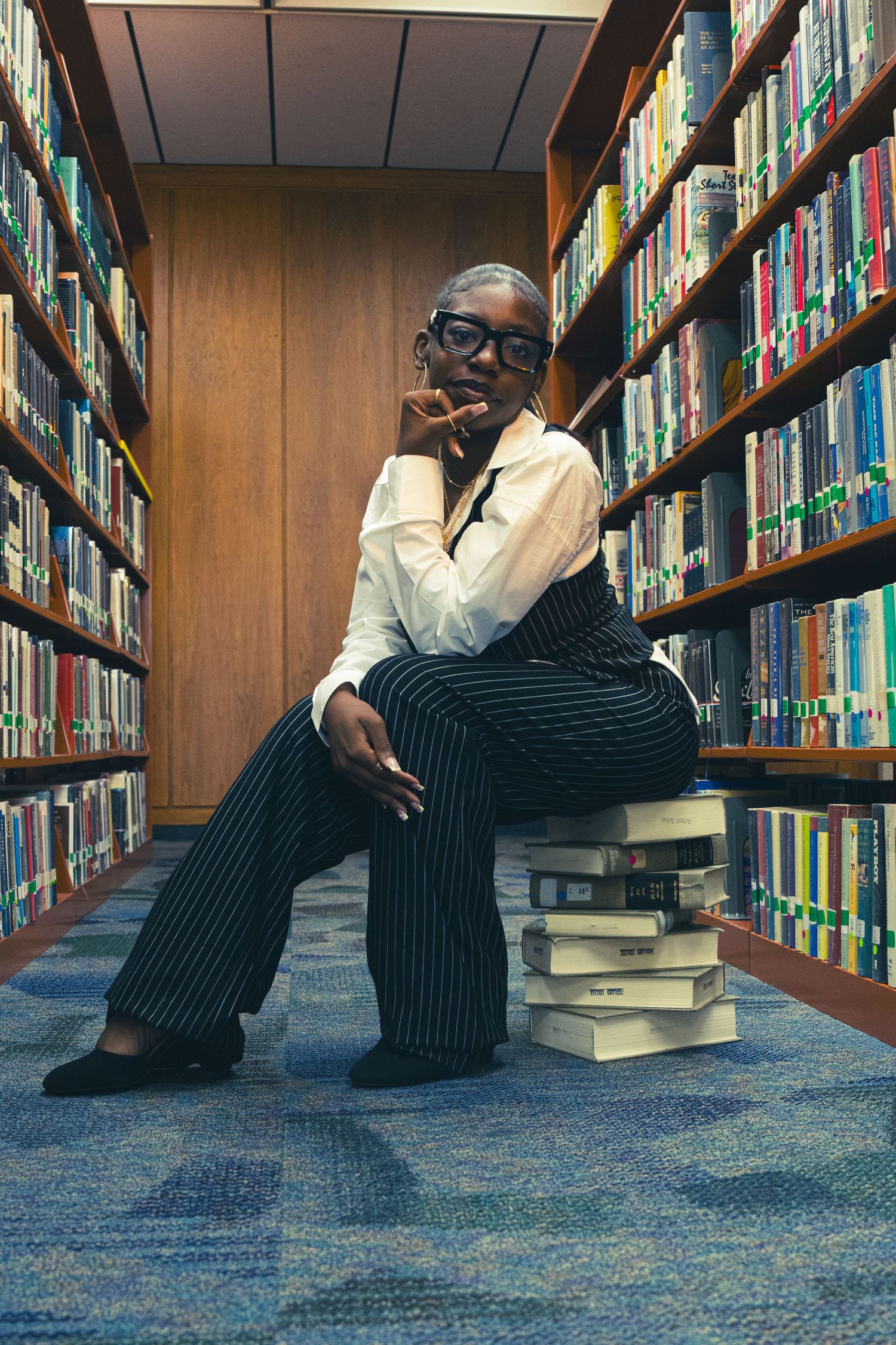 A stylish person in striped pants sits on stacked books in a library aisle, wearing glasses and a white shirt.