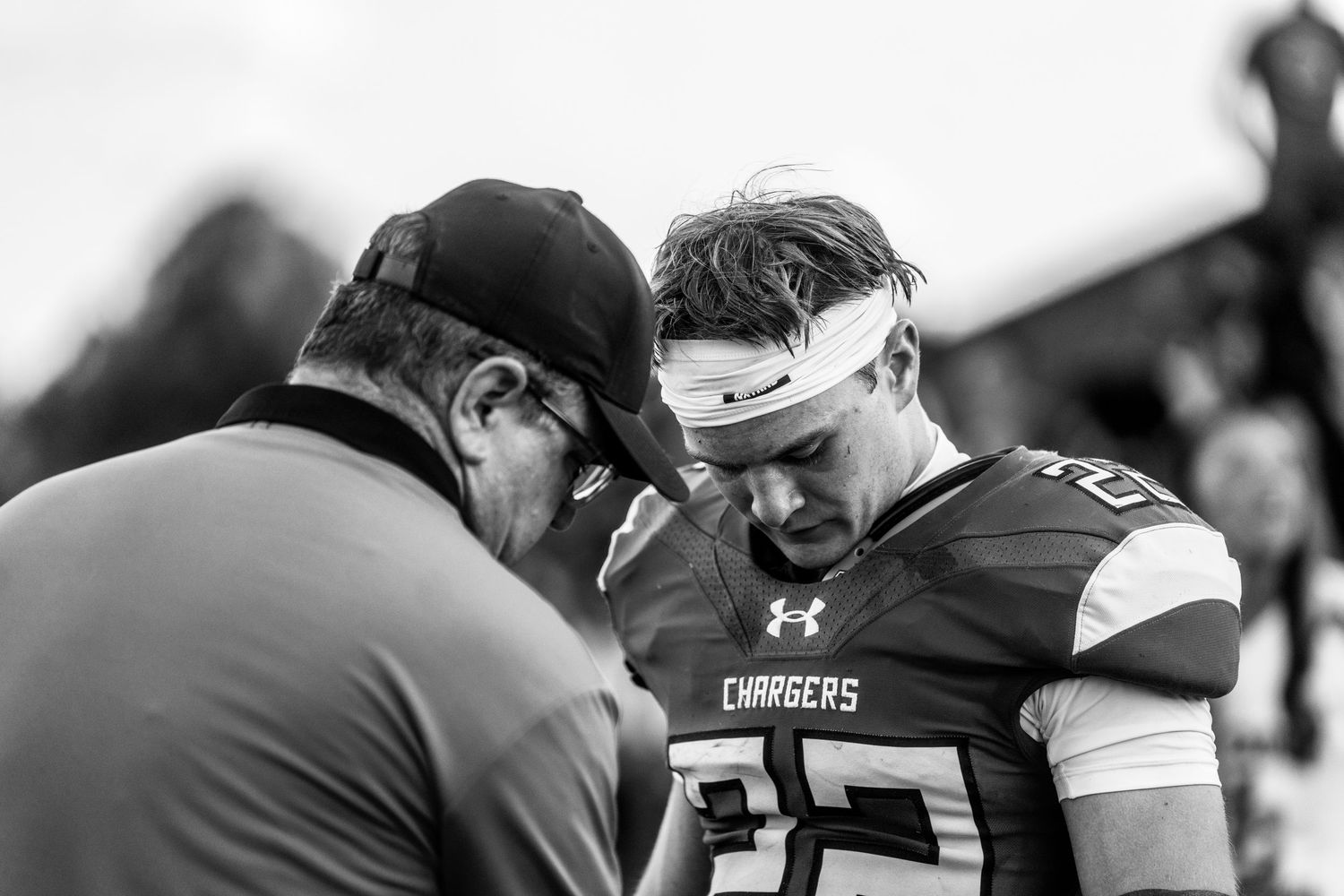 Black and white photo of a Chargers football player with a head bandage receiving coaching on the sideline.