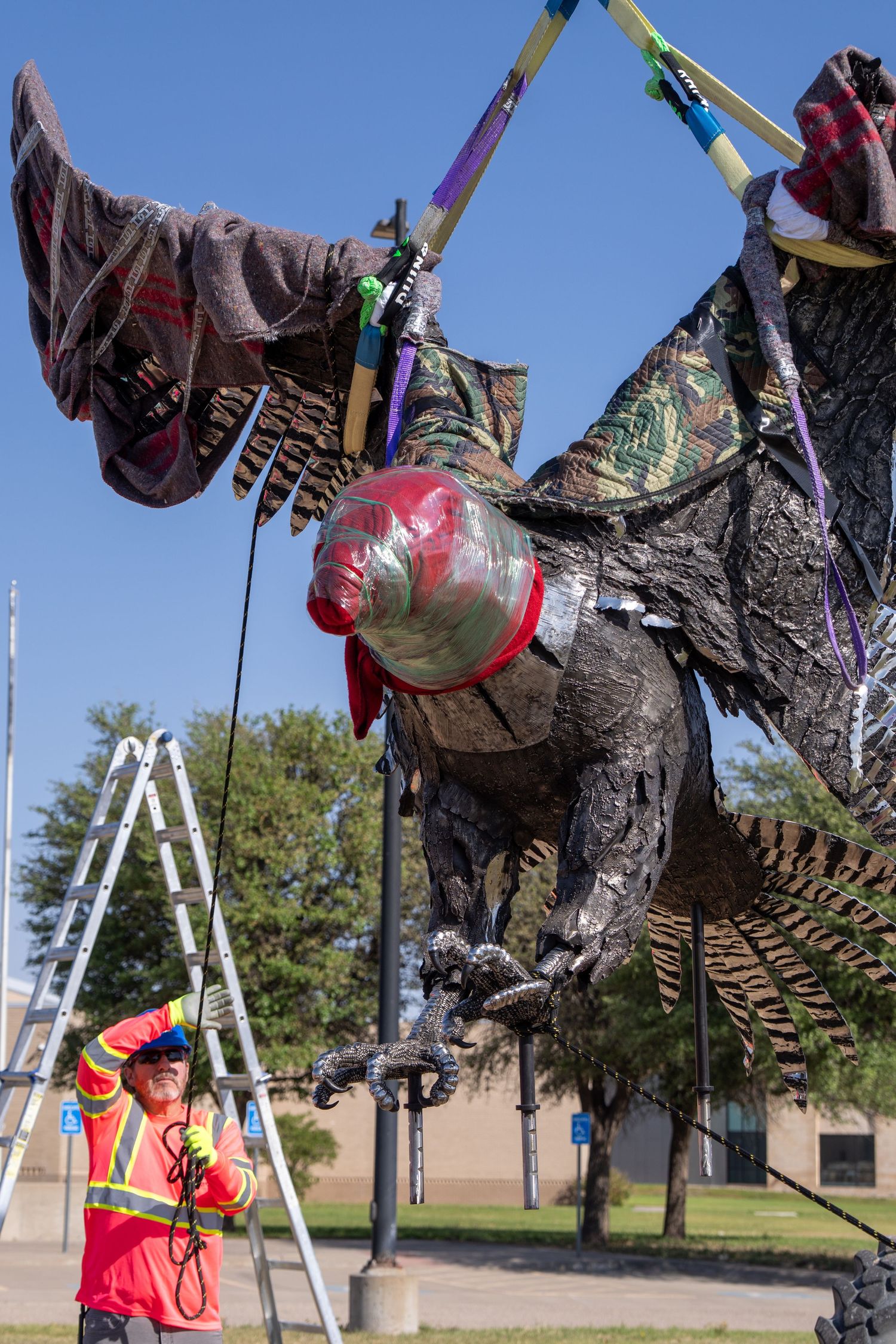 Worker in orange vest guides installation of large dragon sculpture suspended by colorful straps under blue sky.