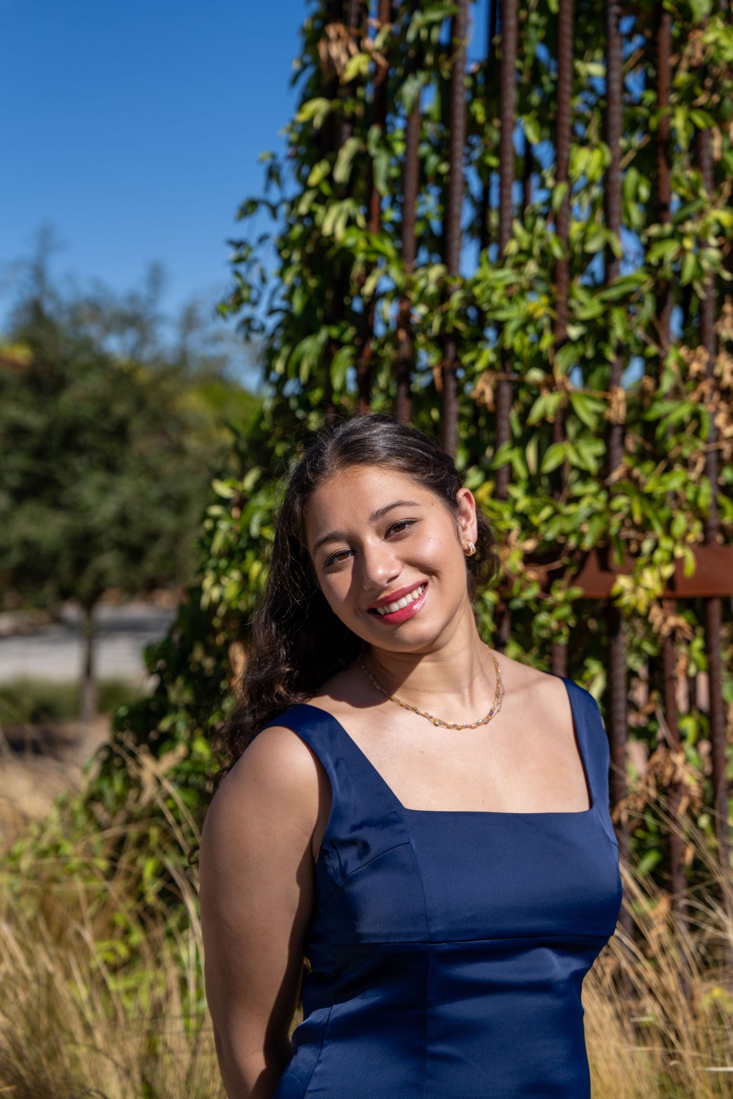 Young woman in blue dress smiling outdoors near ivy-covered rustic metal trellis on a sunny day.
