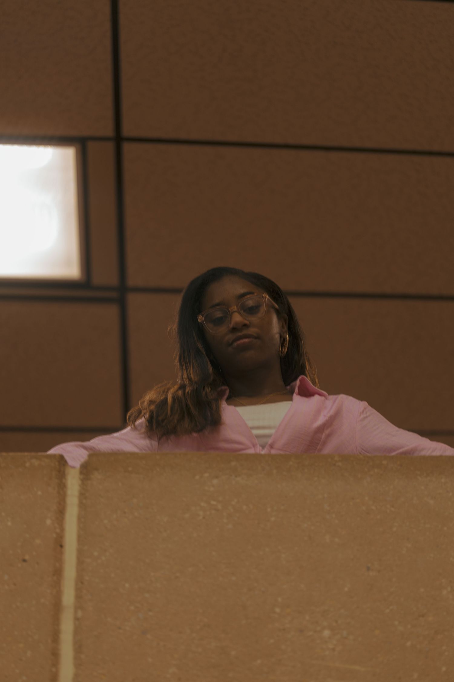 A young Black woman in a pink shirt and round glasses leans on a concrete ledge, looking up with a calm expression.