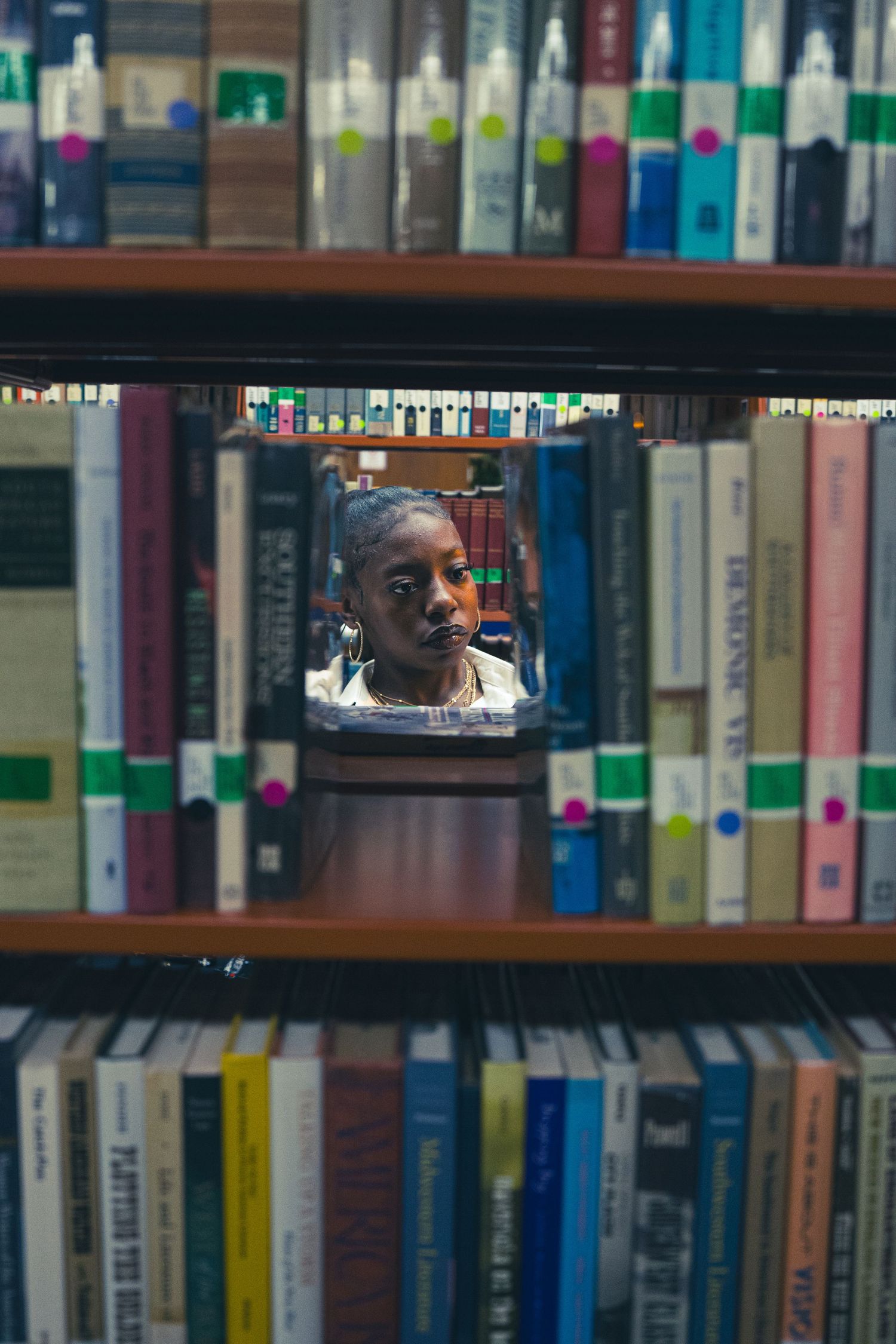 Creative shot of Black woman seen through gap in library bookshelf between colorful books.