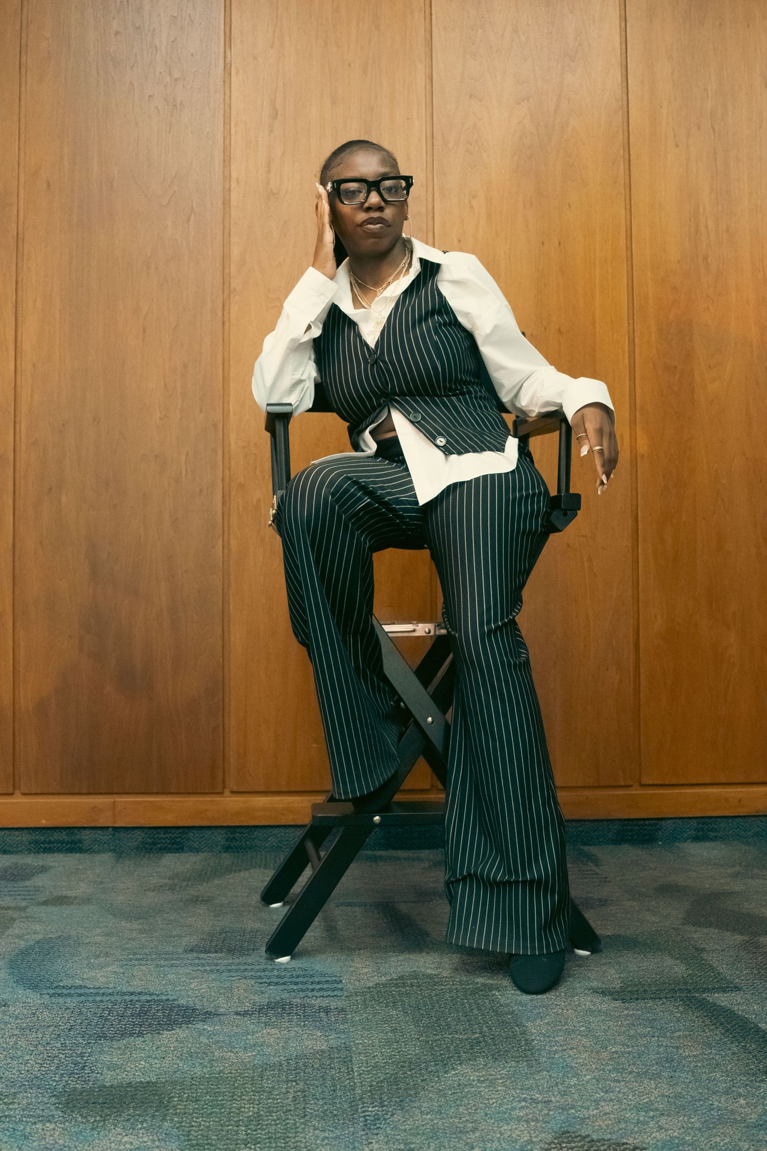 Stylish Black woman in pinstripe suit sits relaxed in a directors chair against a wood-paneled wall.