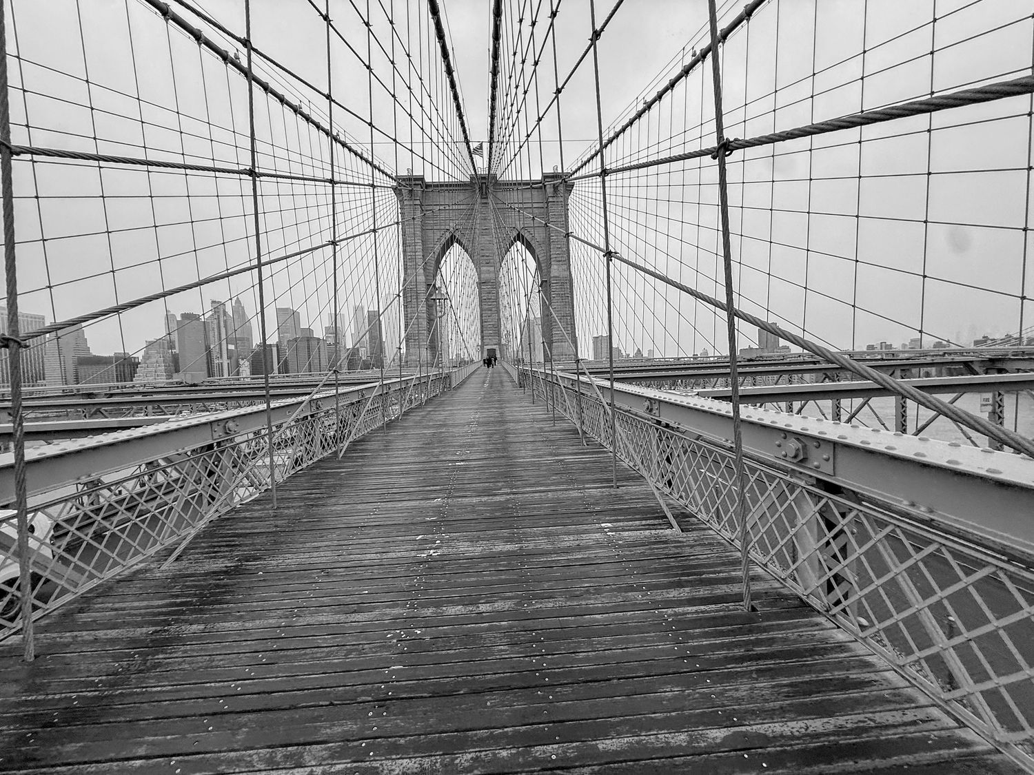 Black and white photo of Brooklyn Bridge walkway with suspension cables and NYC skyline in the background.